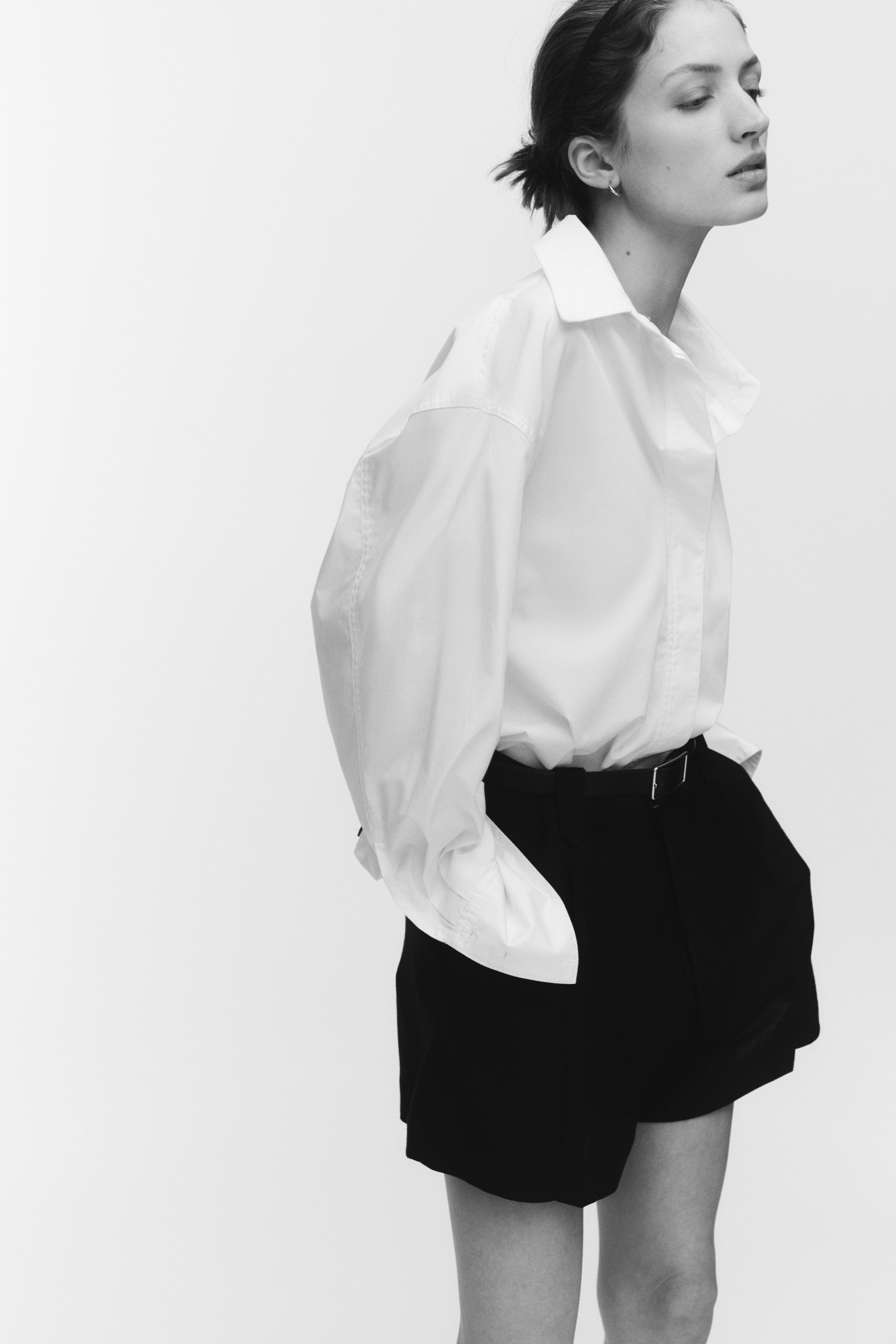 View larger image: Loose-fit na itim na shorts sa isang textured weave ang suot ng modelo, na may mataas na baywang, pahilis na bulsa sa gilid, knife pleats, at mga tupi sa harap. Ang modelo ay nakatuck-in ng puting collared na shirt sa shorts, at isang sinturon na may nakikitang buckle ang nag-aayos sa baywang.
