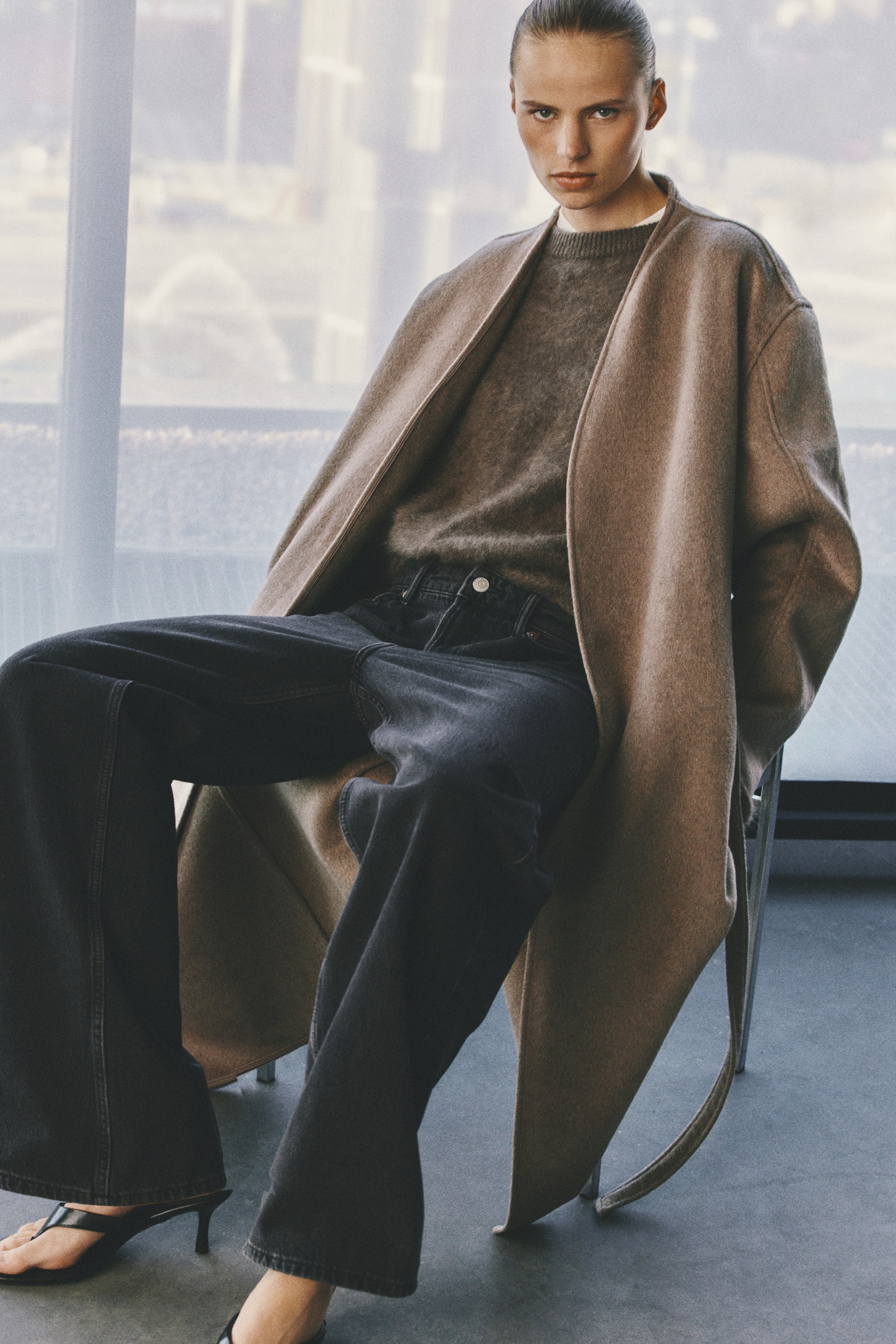 Visualizza immagine più grande: Un cappotto lungo in misto lana infeltrita color marrone chiaro è indossato aperto dalla modella seduta. Il cappotto presenta spalle scese e una cintura da annodare morbida che pende lateralmente, ed è abbinato a un maglione girocollo grigio e jeans grigio scuro.