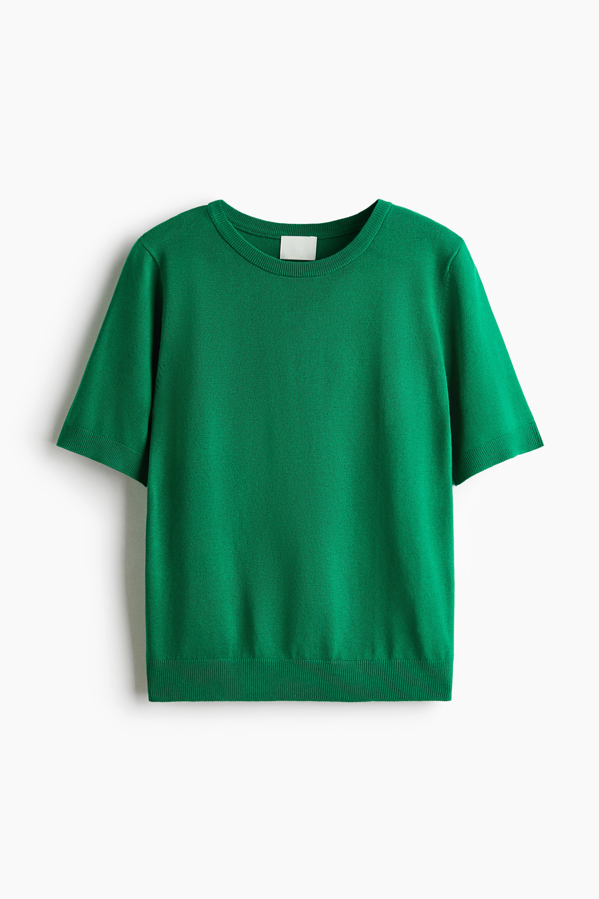 La modelo lleva puesto un suéter de tejido fino verde de manga corta, con ribete blanco en contraste en el escote redondo y los puños. El suéter está metido dentro de unos jeans azules de pierna ancha ajustados con un cinturón negro, y se aprecia una capa de camiseta blanca en el cuello.