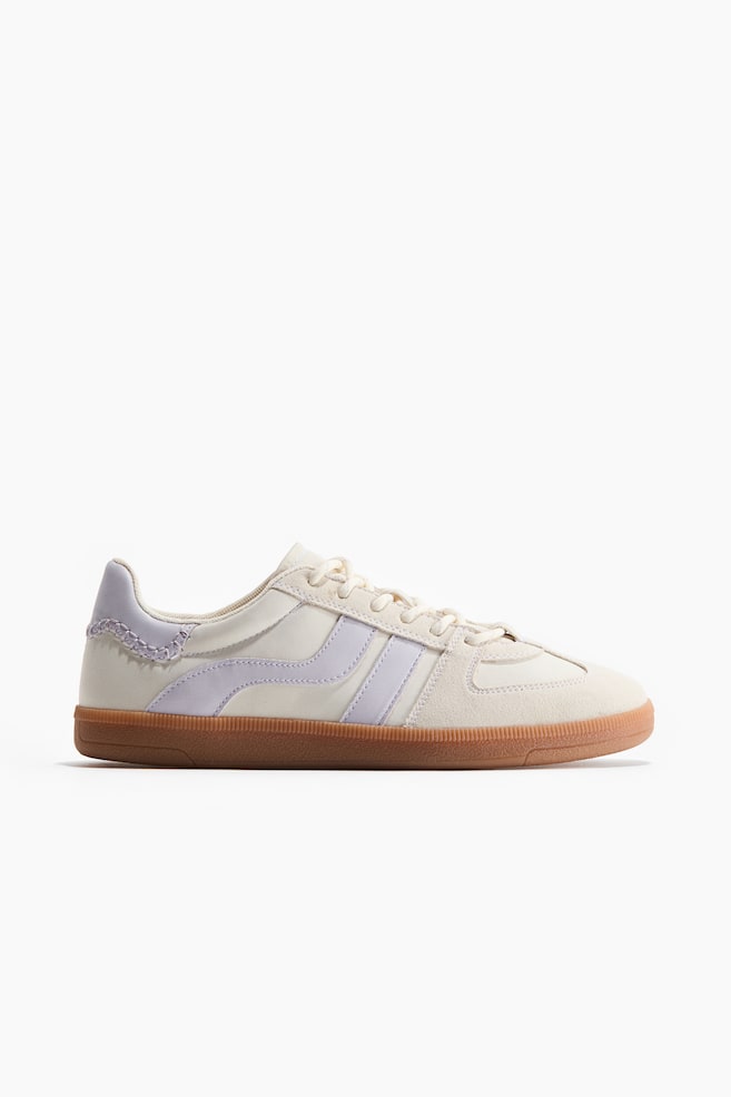 A low-profile trainer features a cream upper with light purple side stripes and a matching heel overlay with visible stitching. It displays cream lacing at the front, a padded top edge, and a brown sole measuring 2.5 cm in height. - 1