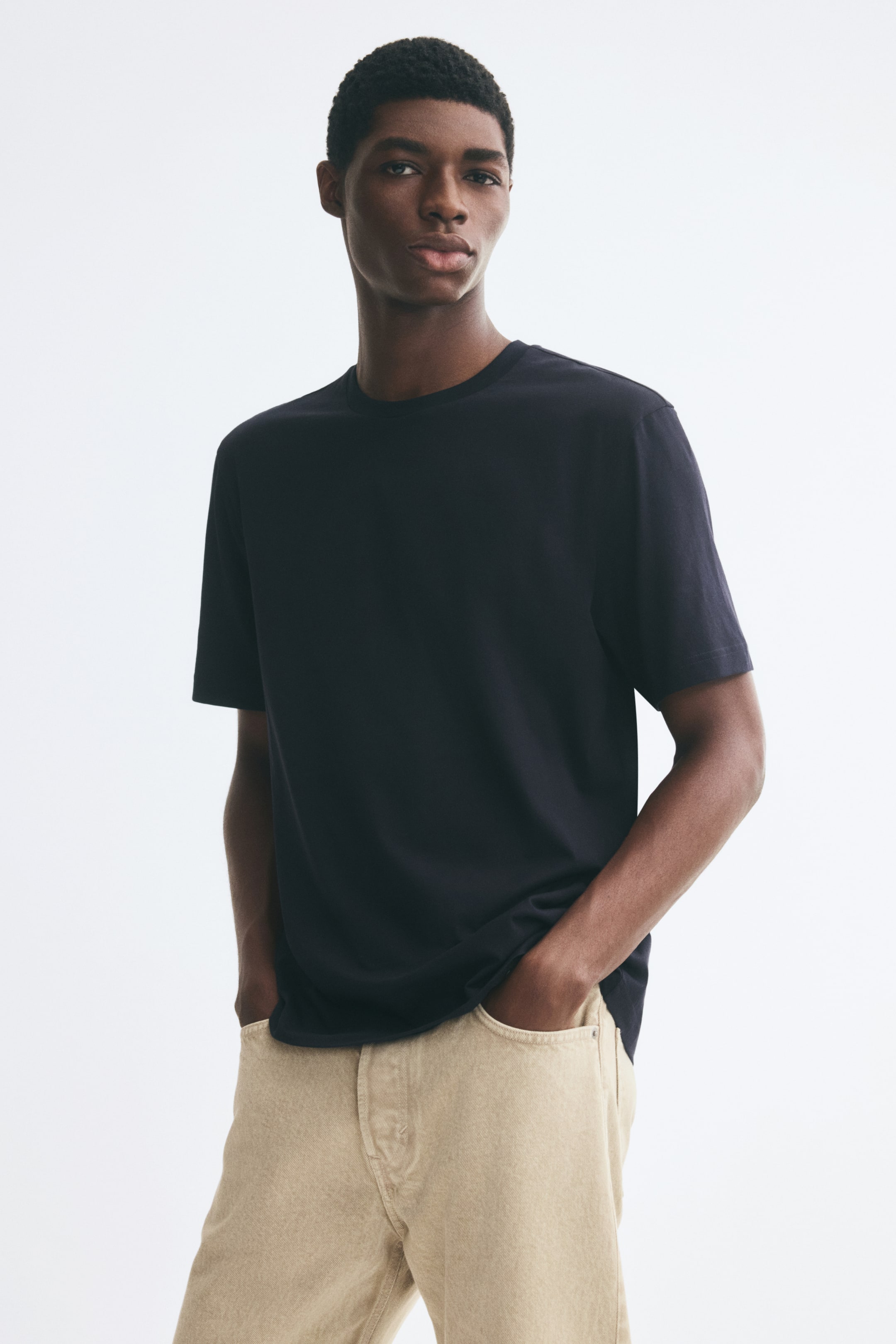 Größeres Bild anzeigen: Ein dunkel marineblaues T-Shirt mit kurzen Ärmeln und Rundhalsausschnitt wird vom Model getragen, das mit den Händen in den Taschen einer beigefarbenen Hose steht. Das T-Shirt verfügt über eine lockere Passform und eine glatte, klare Textur.