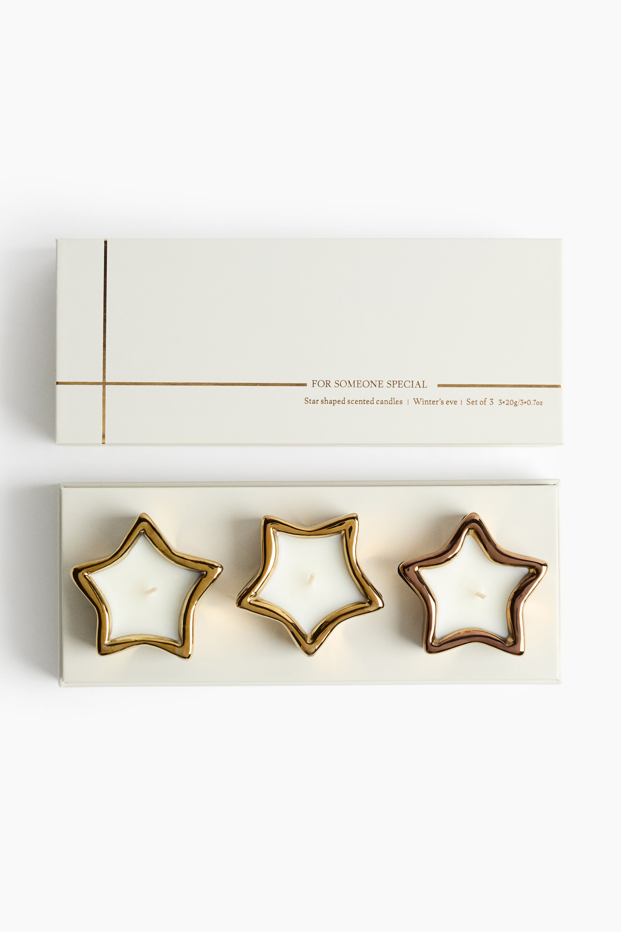 View larger image: A gift set of three star-shaped scented candles is presented in a cream-coloured gift box, shown from a top-down perspective. The candles feature white wax and single wicks housed in metallic star-shaped holders with gold and bronze-toned finishes. The matching lid displays minimalist gold line details and elegant text.