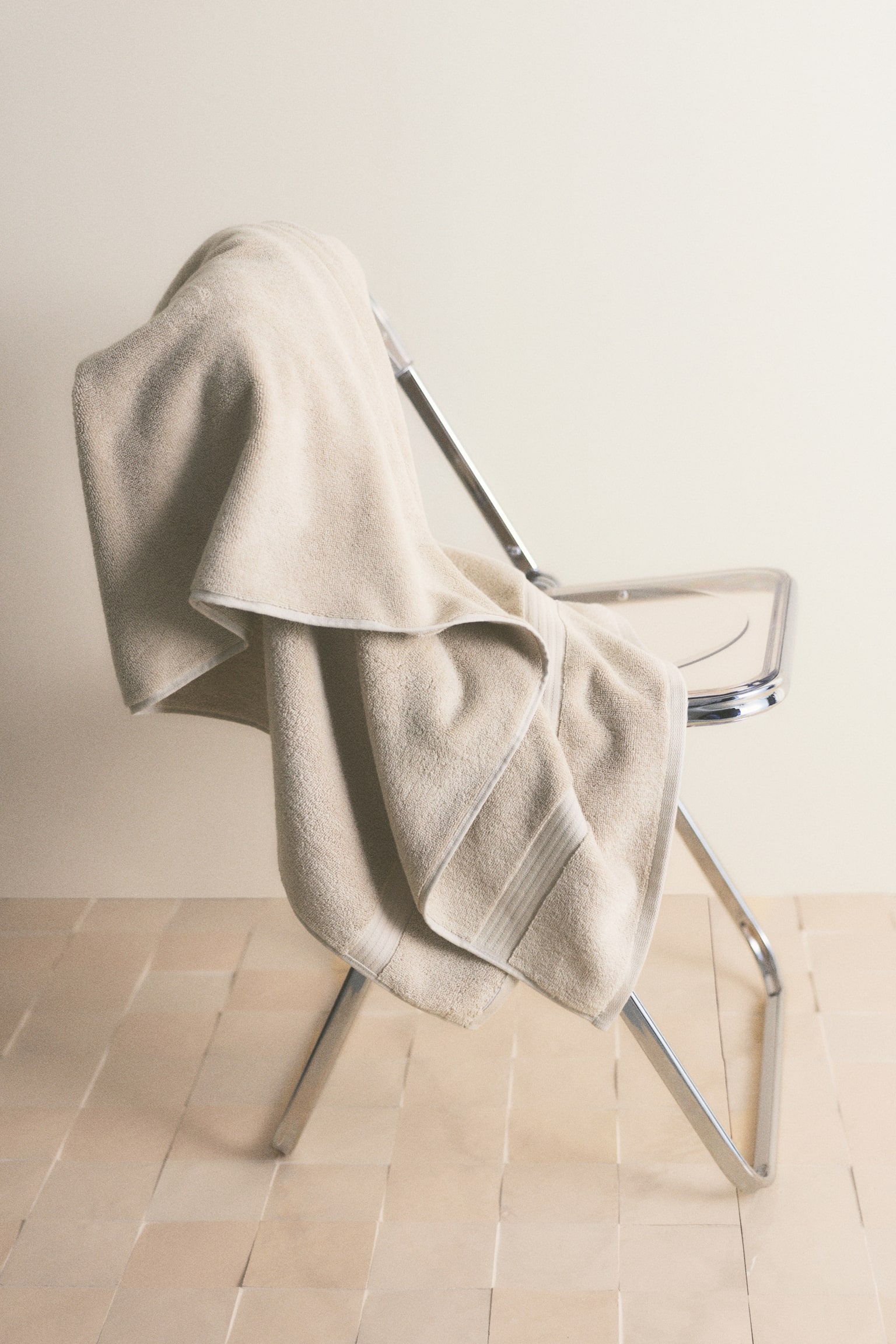 A light beige towel hangs from a hook, showcasing a soft texture and a subtle horizontal decorative band. It drapes with gentle folds, with one part overlapping another. - 1