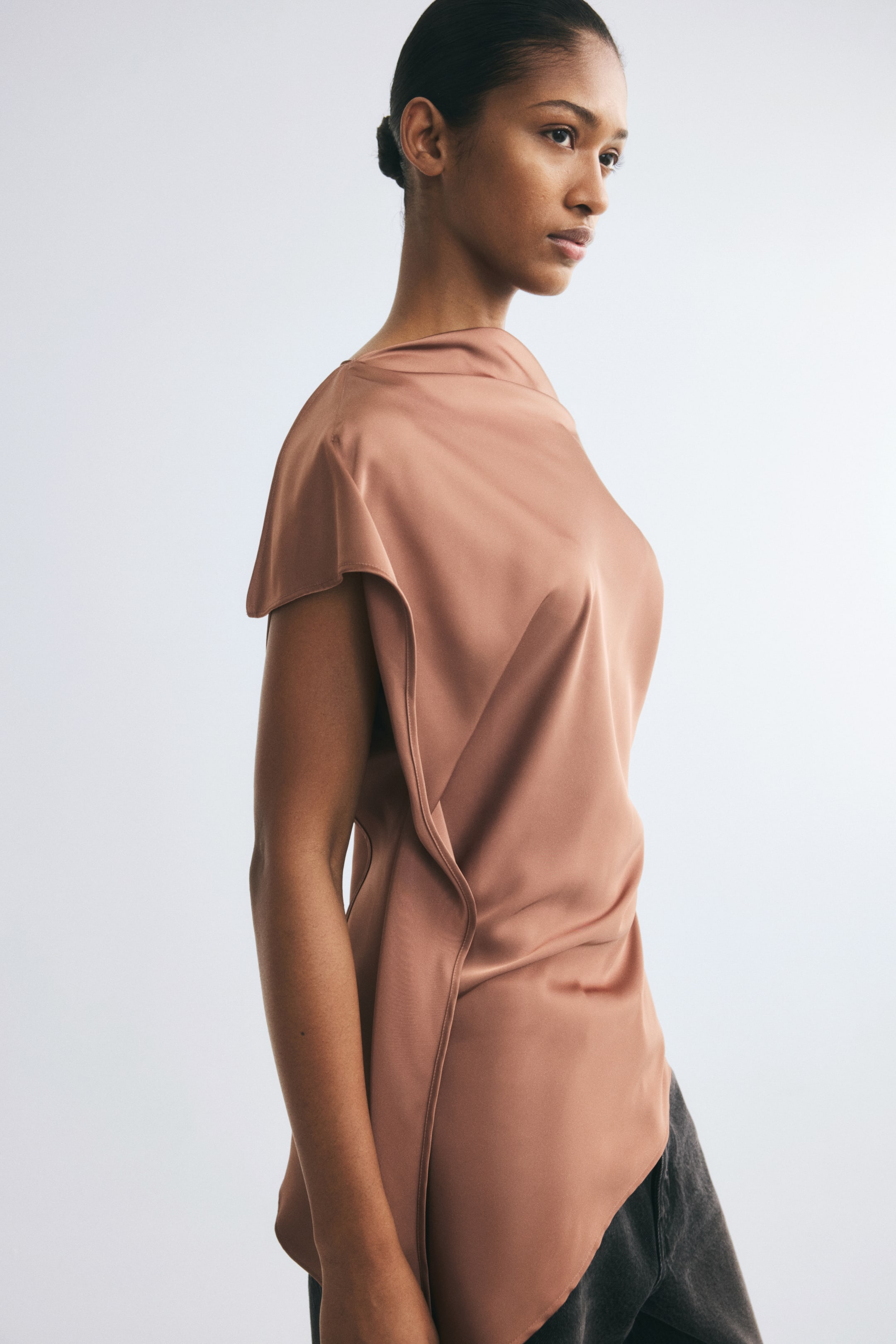Ampliar la imagen: La modelo lleva puesta una blusa de satén color rosa, con escote asimétrico, mangas cortas tipo cap sleeve y paneles drapeados en un costado. La blusa tiene un dobladillo asimétrico y se combina con pantalones gris oscuro.