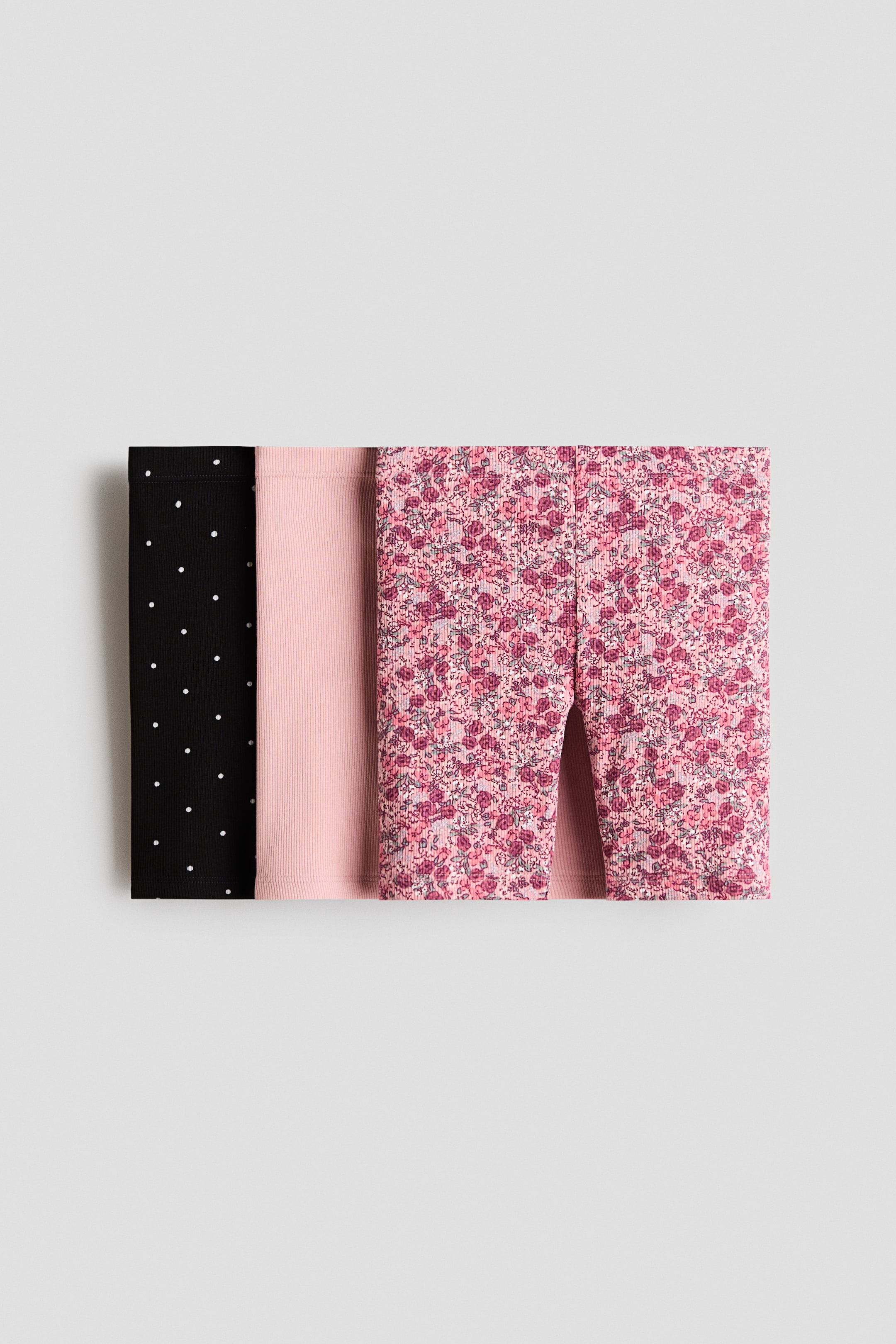 View larger image: Three pairs of ribbed leggings are laid out flat, folded side by side. The leftmost pair is black with white polka dots, the middle pair is a solid light pink, and the rightmost pair is pink with a multicolored floral pattern.