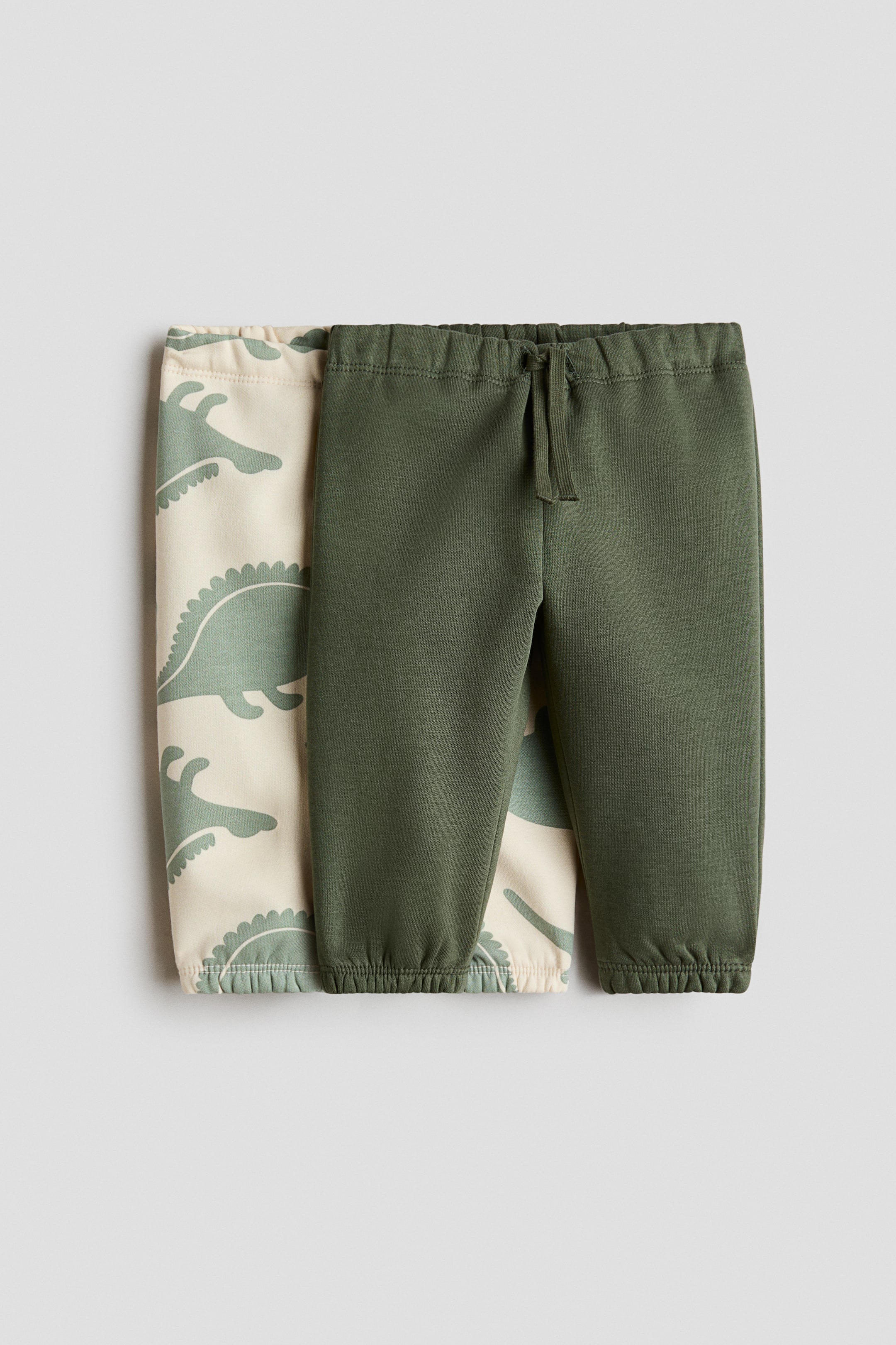 View larger image: Two pairs of joggers are shown from the front, with an olive green pair layered over a cream pair featuring a repeating green dinosaur print. The olive green joggers feature an elasticated waist with a decorative drawstring and covered elastication at the hems, while the cream pair also features covered elastication at the hems.