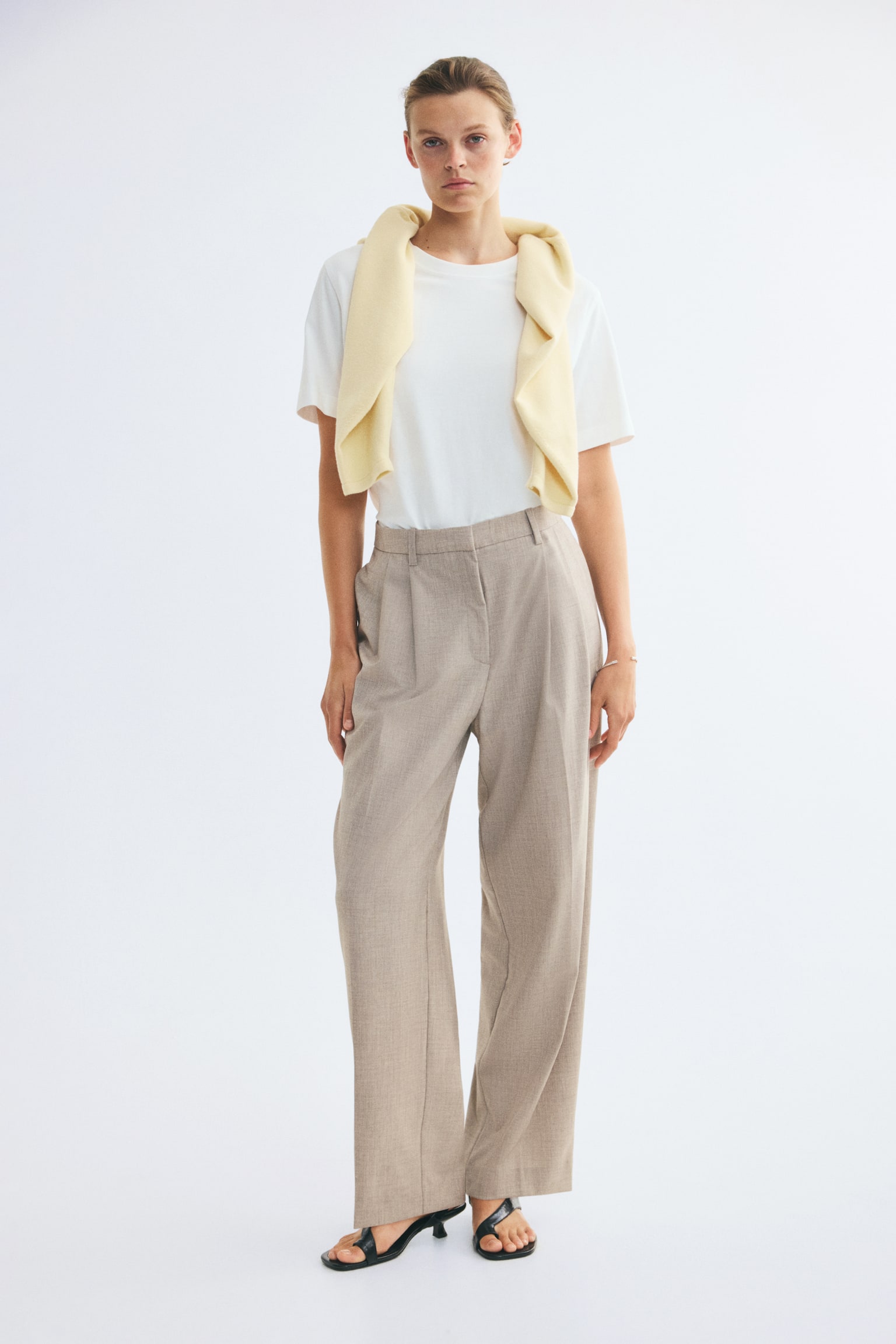 Trousers feature a light brown, subtly textured fabric, a flat waistband with belt loops, and prominent front pleats, presenting a relaxed, straight-leg style from a front-facing view. - 1
