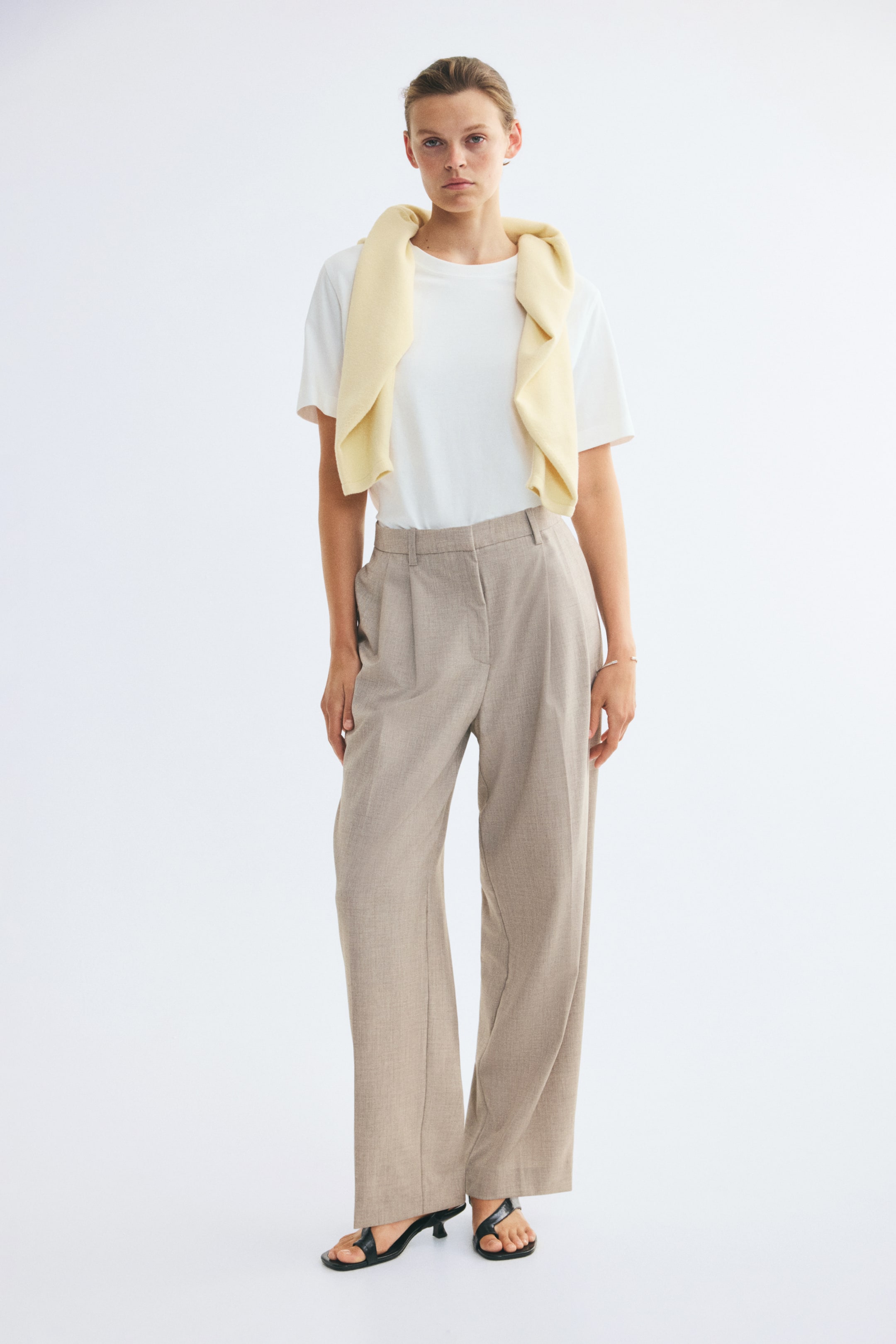View larger image: Tailored trousers in a light beige woven fabric are being worn by the model, featuring a high-waisted fit, belt loops, front pleats, and creases down the wide legs. The trousers are styled with a tucked-in white T-shirt and a light yellow sweater draped over the shoulders.