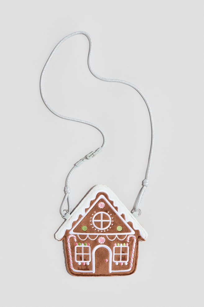 A gingerbread house-shaped bag is facing front, showcasing a brown felt-like body with white plush trim along the roof. The bag is displaying embroidered white icing outlines, windows, a door, and colorful candy designs, and is hanging from a thin, silver cord strap. - 1