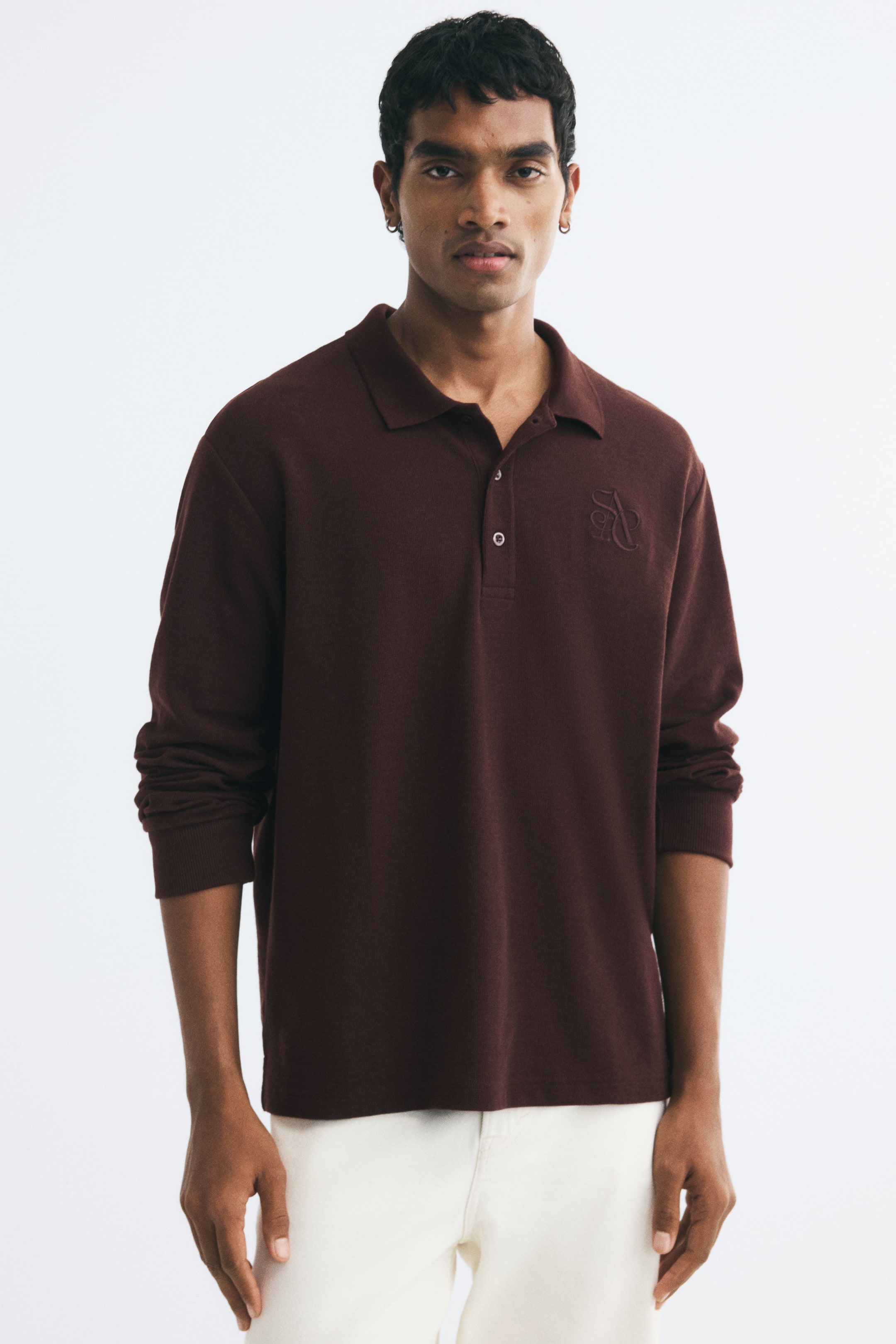 View larger image: A dark brown, long-sleeved polo shirt in waffle-knit fabric is being worn by the model. The shirt features a ribbed collar, a button placket, and a tonal embroidered detail on the chest, and is paired with off-white trousers.