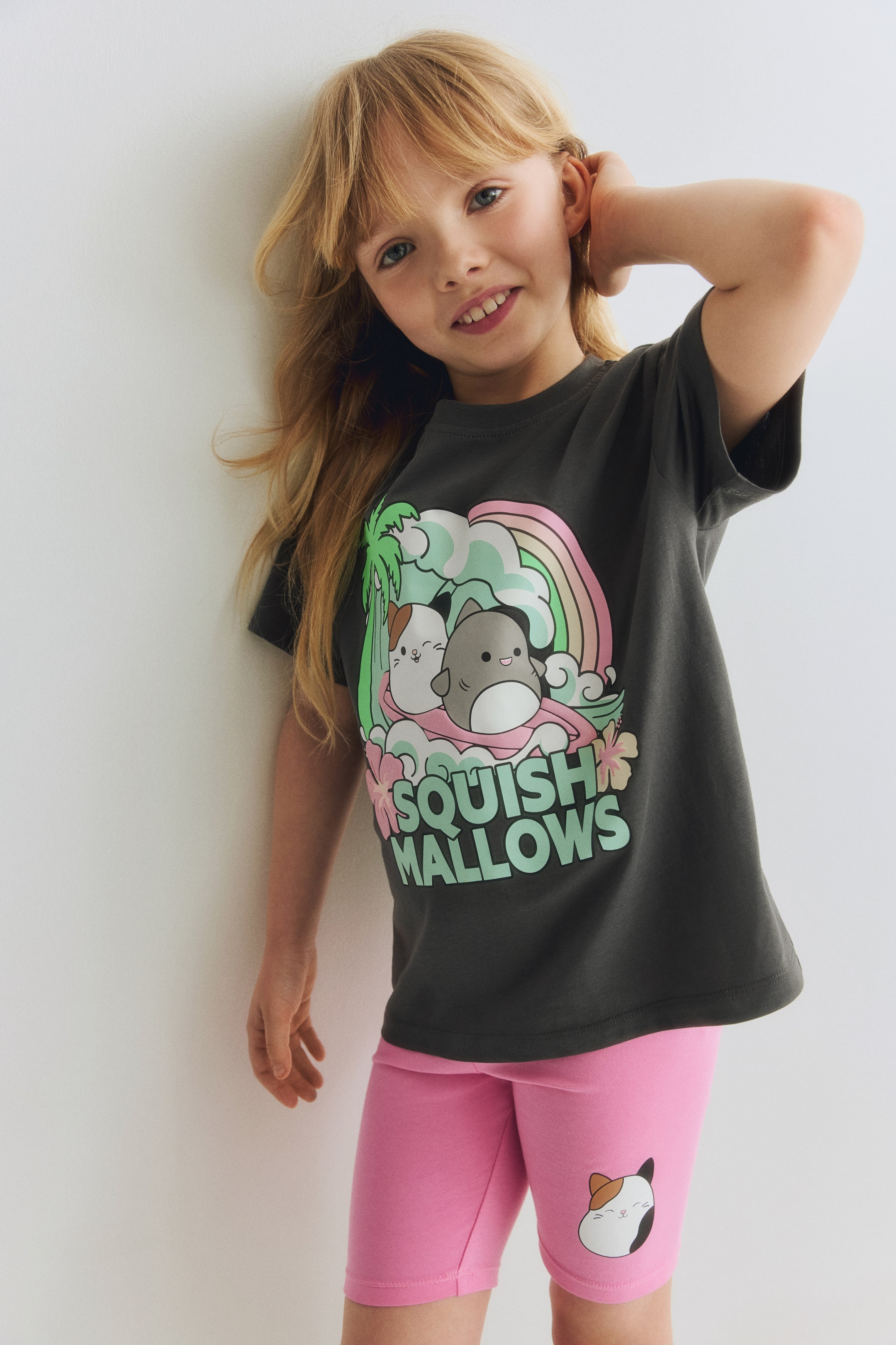 View larger image: A dark grey short-sleeved t-shirt and pink cycling shorts are being showcased, angled slightly to the right. The t-shirt features a vibrant graphic of two Squishmallow characters on a pink wave, accompanied by a rainbow and tropical plants, above the text 'SQUISHMALLOWS'. The shorts display a small calico cat head graphic on the lower left leg.