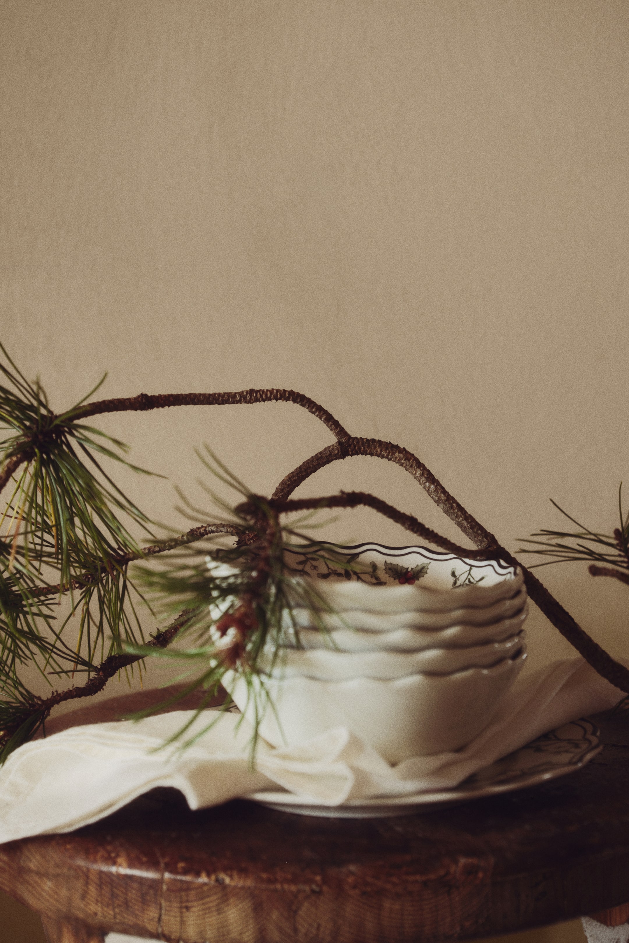 Voir une image plus grande: Des bols en porcelaine blanche, orientés vers l’avant, sont empilés sur un tissu blanc. Ils présentent des bords ondulés avec des contours accentués foncés et un motif festif de houx à l’intérieur du bol du dessus. Une branche de pin décorative est drapée sur la pile, mettant en valeur leur design festif.