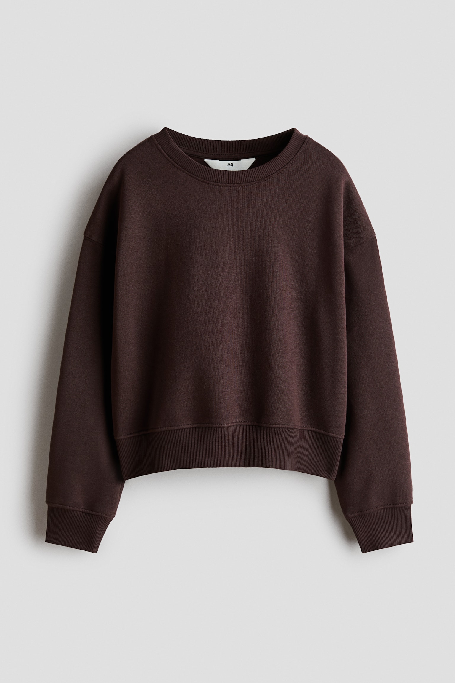 A dark brown, loose-fit sweatshirt is shown from the front, featuring a round ribbed neckline, dropped shoulders, and long sleeves. The garment presents a relaxed, boxy silhouette with thick ribbed cuffs and a matching ribbed hem.
