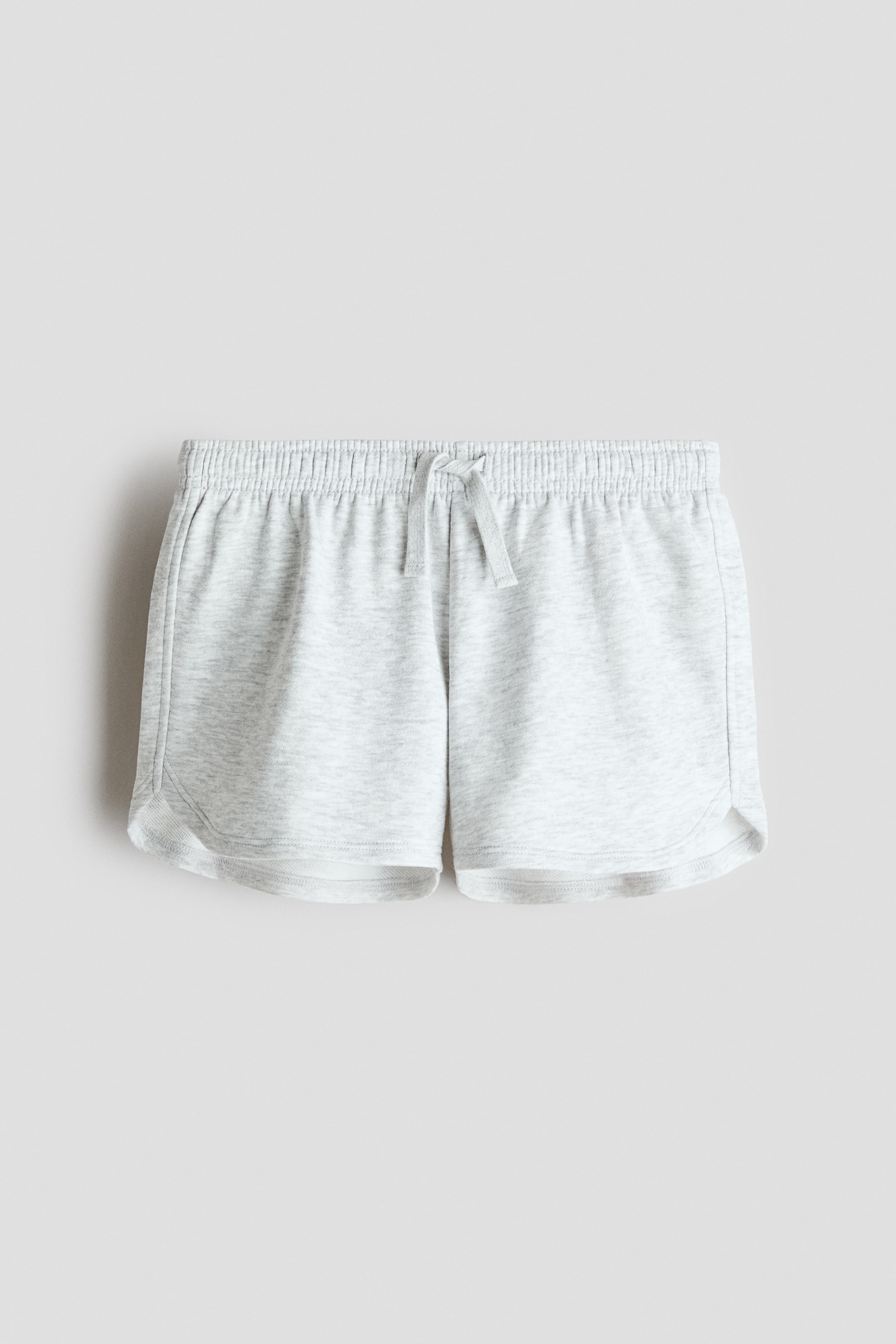 Xem ảnh lớn hơn: Quần short dáng rộng được làm từ chất liệu vải nỉ màu xám nhạt, nhìn từ phía trước. Chúng có phần eo co giãn với dây rút buộc thành hình nơ, và đường viền cong ở hai bên ngoài.