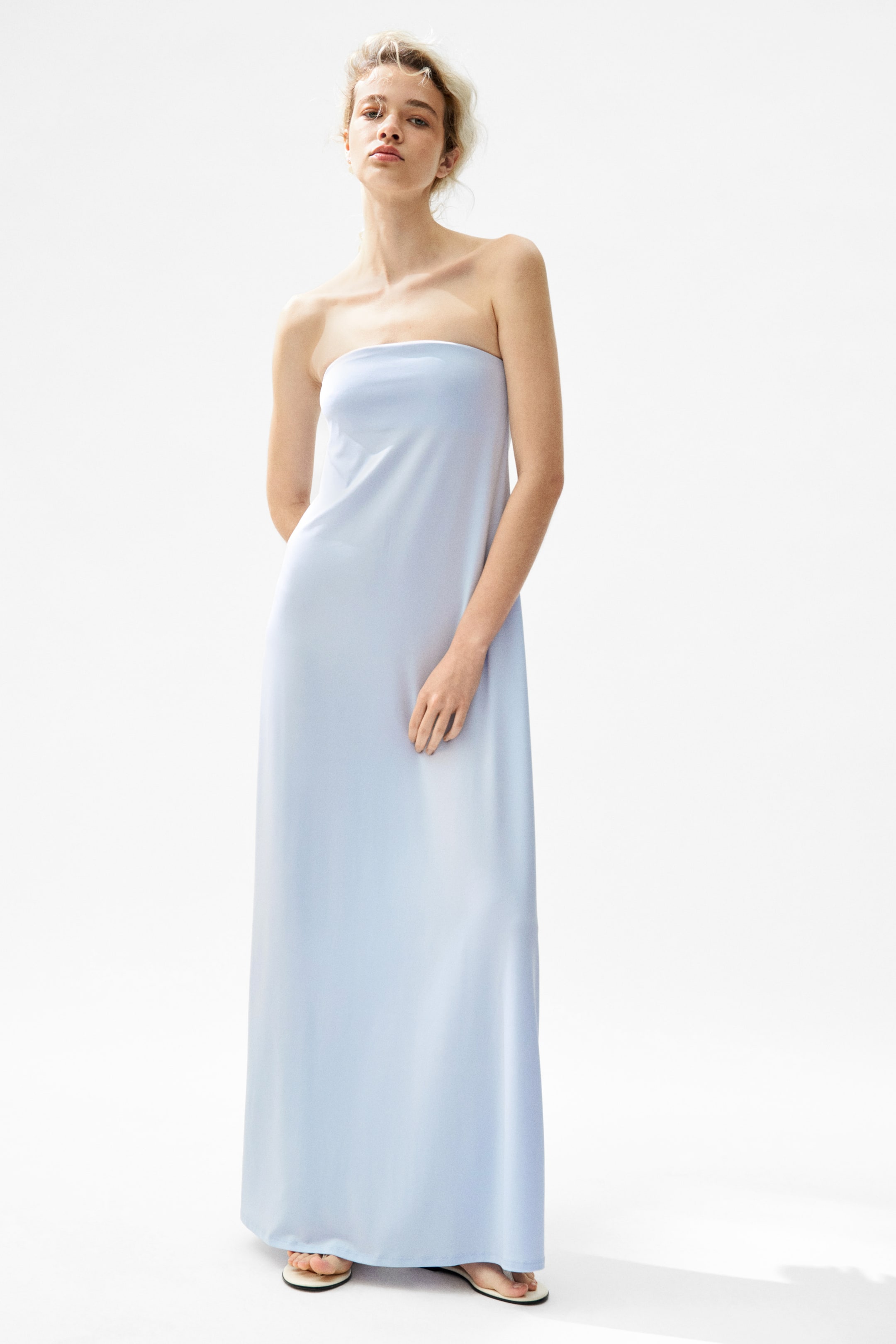View larger image: The model is wearing a light blue strapless maxi dress that showcases a fluid, floor-length silhouette. She completes the look with white flat sandals.