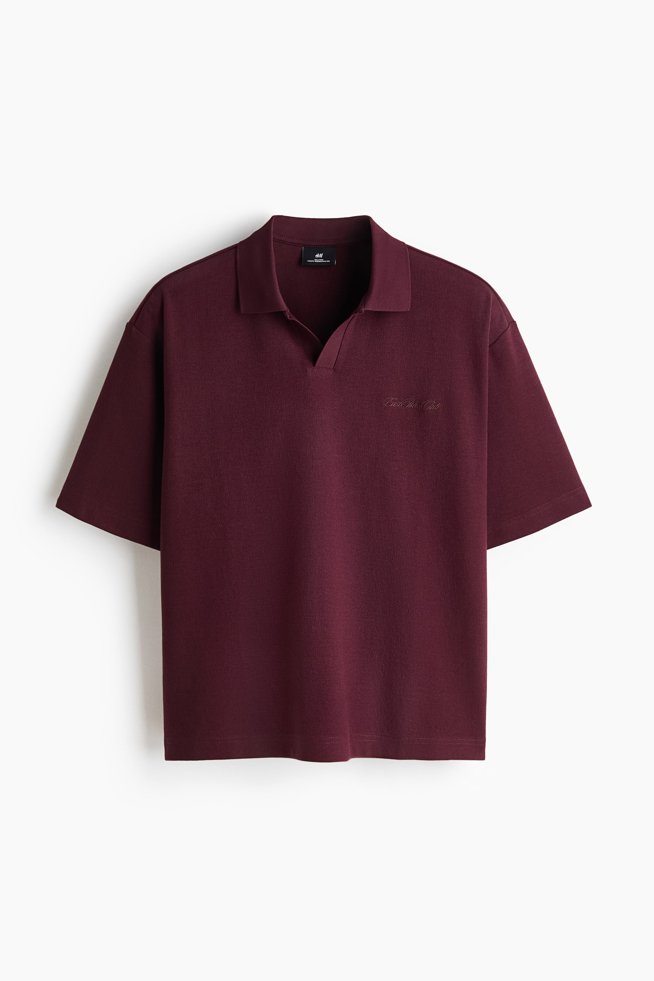 View larger image: A burgundy short-sleeved polo shirt is shown facing front, featuring a buttonless open collar, dropped shoulders, and wide sleeves for a relaxed fit. The garment is constructed with a straight hem and features subtle, tonal script embroidery on the left chest.