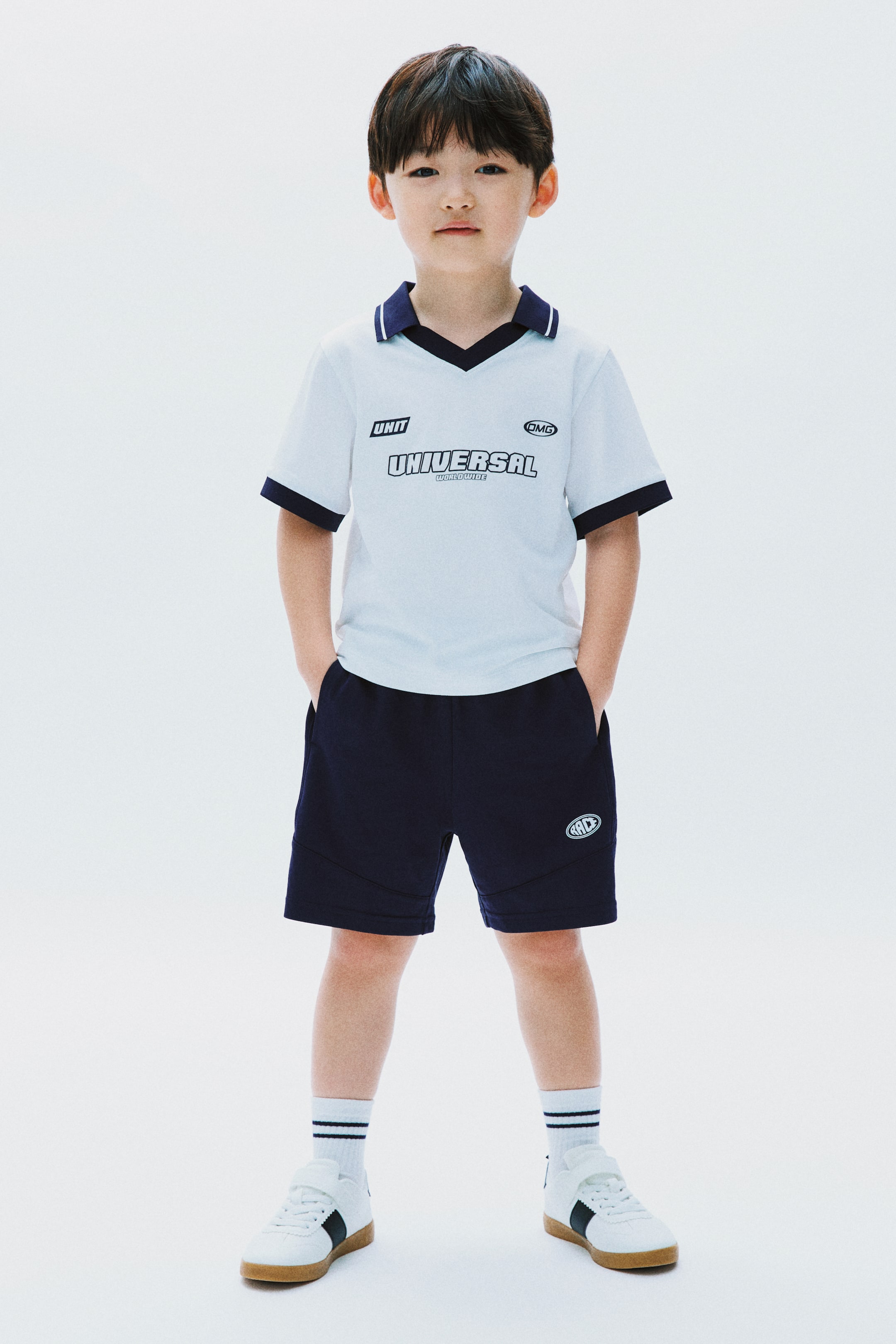 拡大画像を表示: ライトブルーの半袖トップスには、ネイビーのポロ襟と袖口があり、胸元には「UNIT」、「OMG」、「UNIVERSAL WORLDWIDE」の文字が表示されています。ネイビーのショートパンツには、左太ももに白い円形のロゴが特徴で、両方の衣服が正面を向いてセットアップとして紹介されています。
