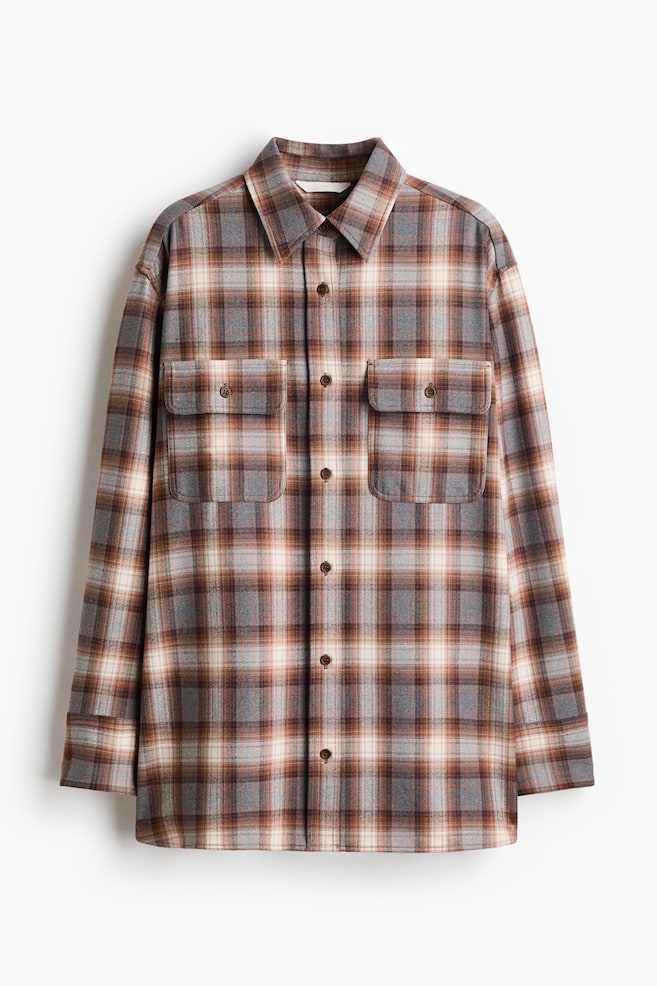 A long-sleeved plaid shirt is hanging facing front, displaying a checkered pattern in shades of brown, grey, and cream. The shirt is featuring a point collar, a button-down front closure, and two symmetrical chest pockets with buttoned flaps. - 1