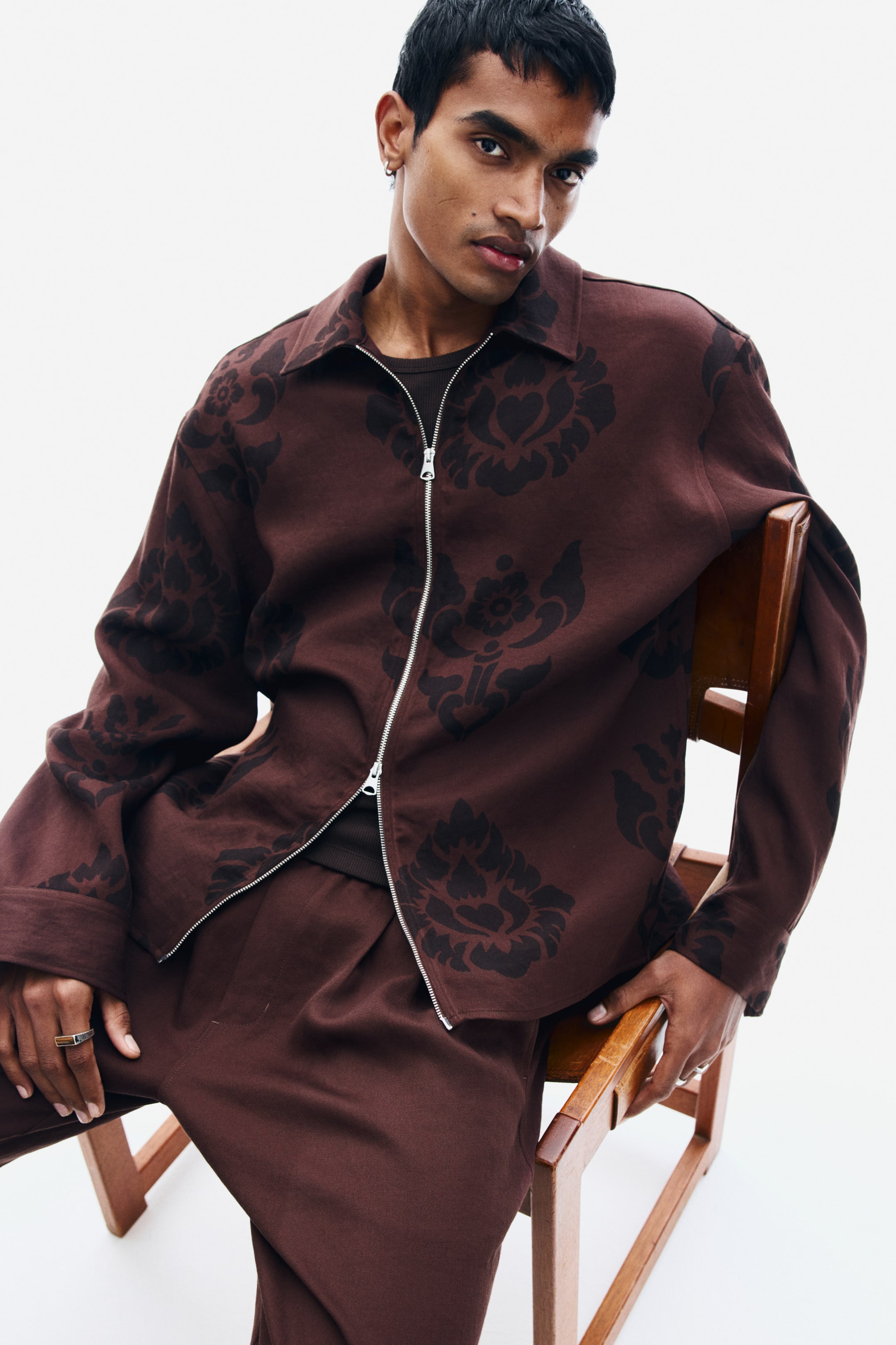 View larger image: A brown twill overshirt featuring a dark damask pattern, a collar, and a silver-toned two-way zipper is being worn by the model. The overshirt is styled partially zipped, layering over a dark brown T-shirt and matching brown trousers.