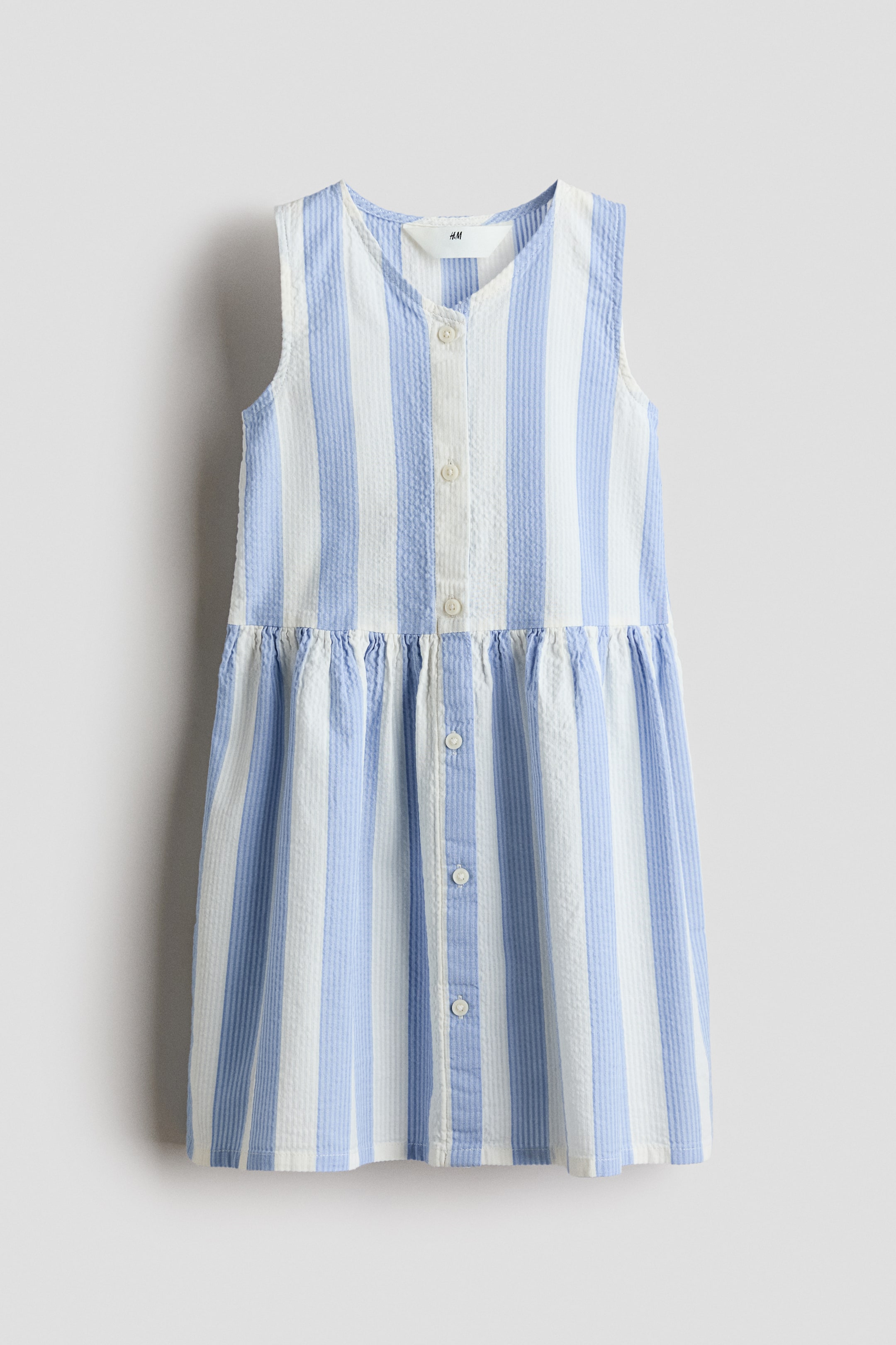 View larger image: Front view of a sleeveless dress, crafted from patterned cotton seersucker in vertical blue and white stripes. It features a V-neck, a gathered seam at the waist leading to a flared skirt, and a button band running down the front, showcasing functional buttons at the top and decorative buttons at the bottom.