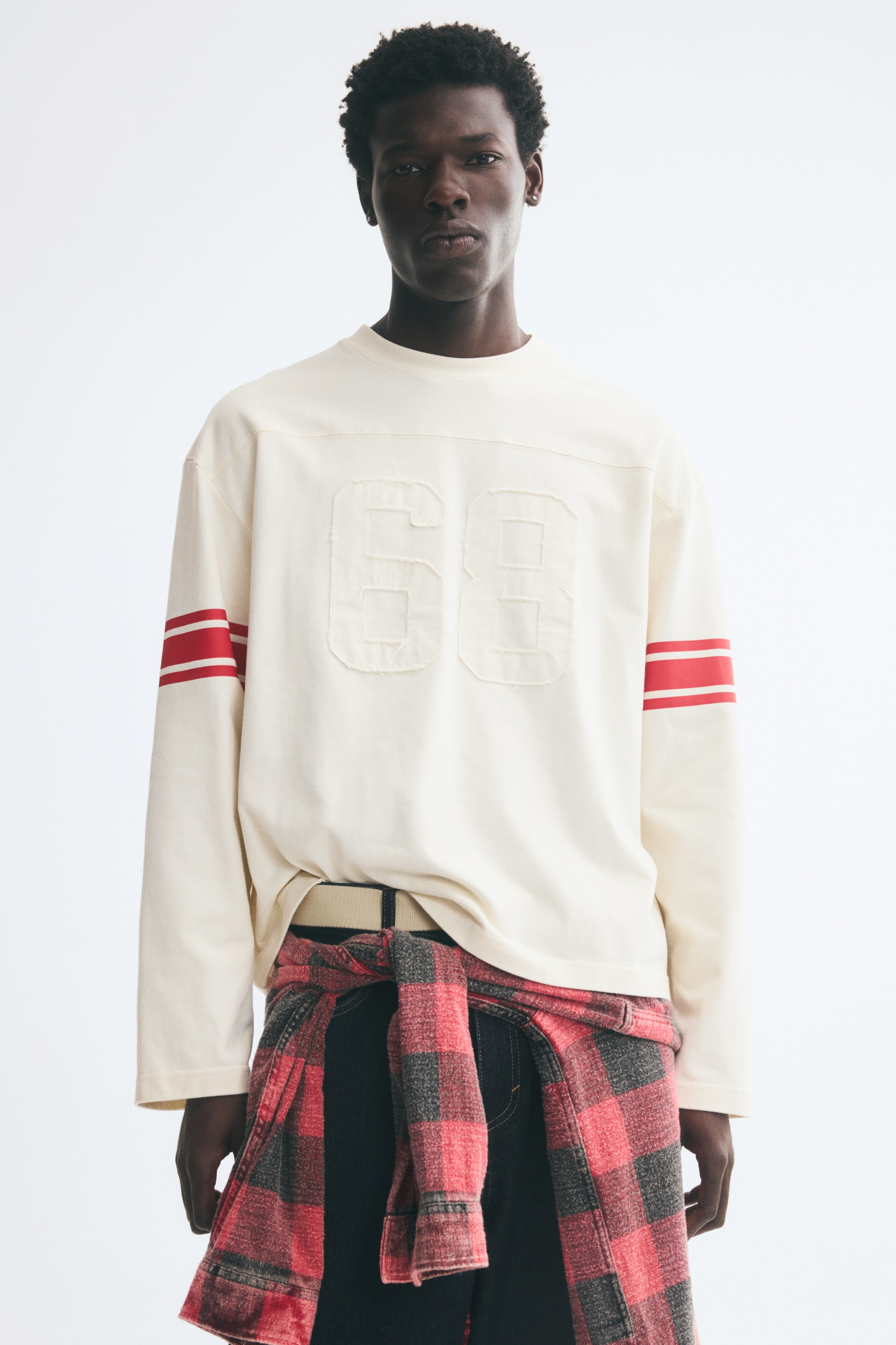 A cream-colored, long-sleeved top is shown facing front, featuring a ribbed crew neck and relaxed drop shoulders. It is displaying a tonal, raw-edged appliquï¿½ of the number "68" on the center chest and two horizontal red stripes on each upper sleeve.