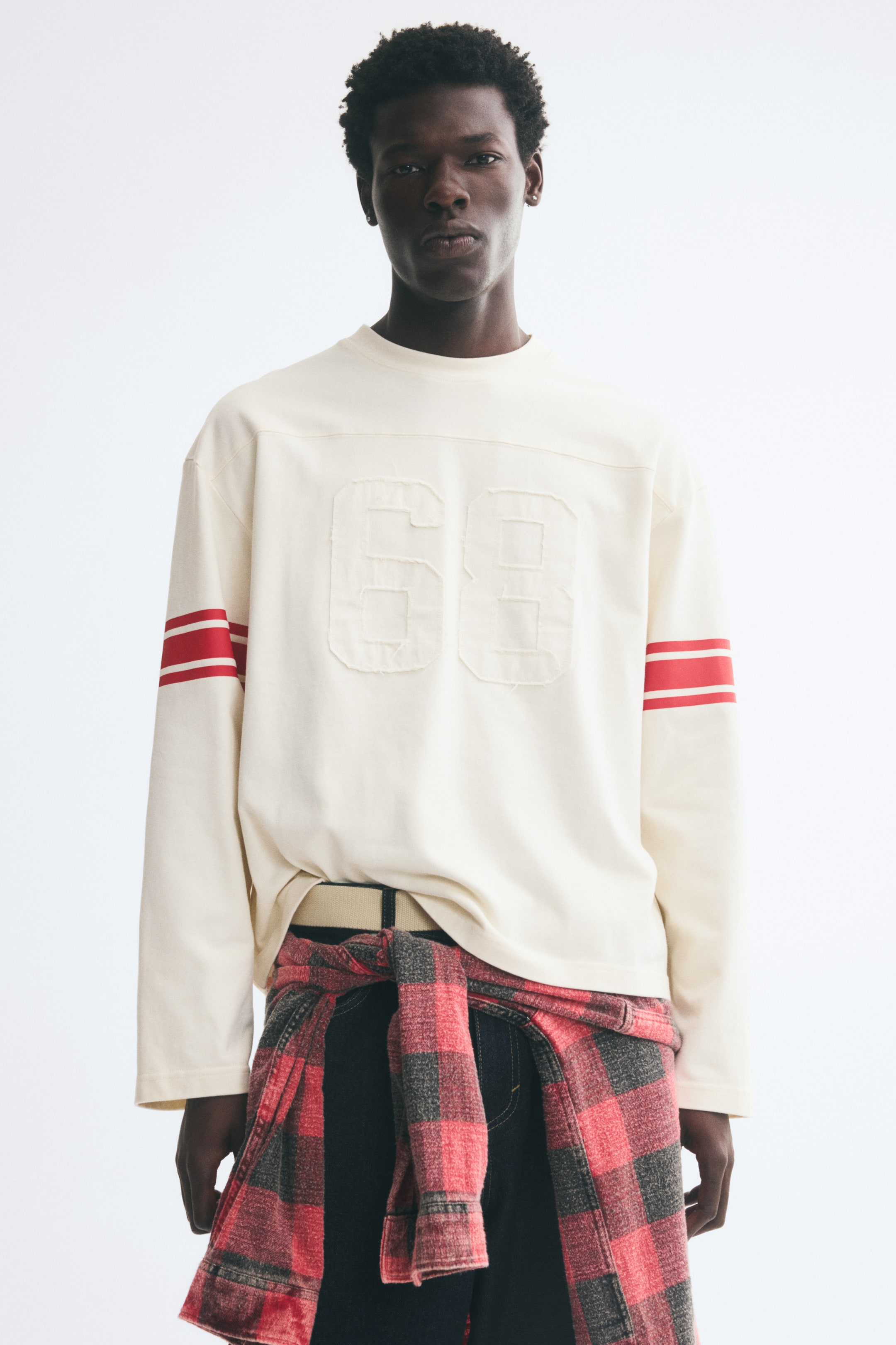 Ampliar la imagen: La modelo lleva puesta una playera color crema de manga larga, con corte relajado, canesú delantero, hombros caídos, un parche "68" de borde sin rematar en el pecho y dobles franjas rojas en las mangas. La playera se estiliza con una camisa de cuadros rojos y negros amarrada a la cintura sobre pantalones oscuros.