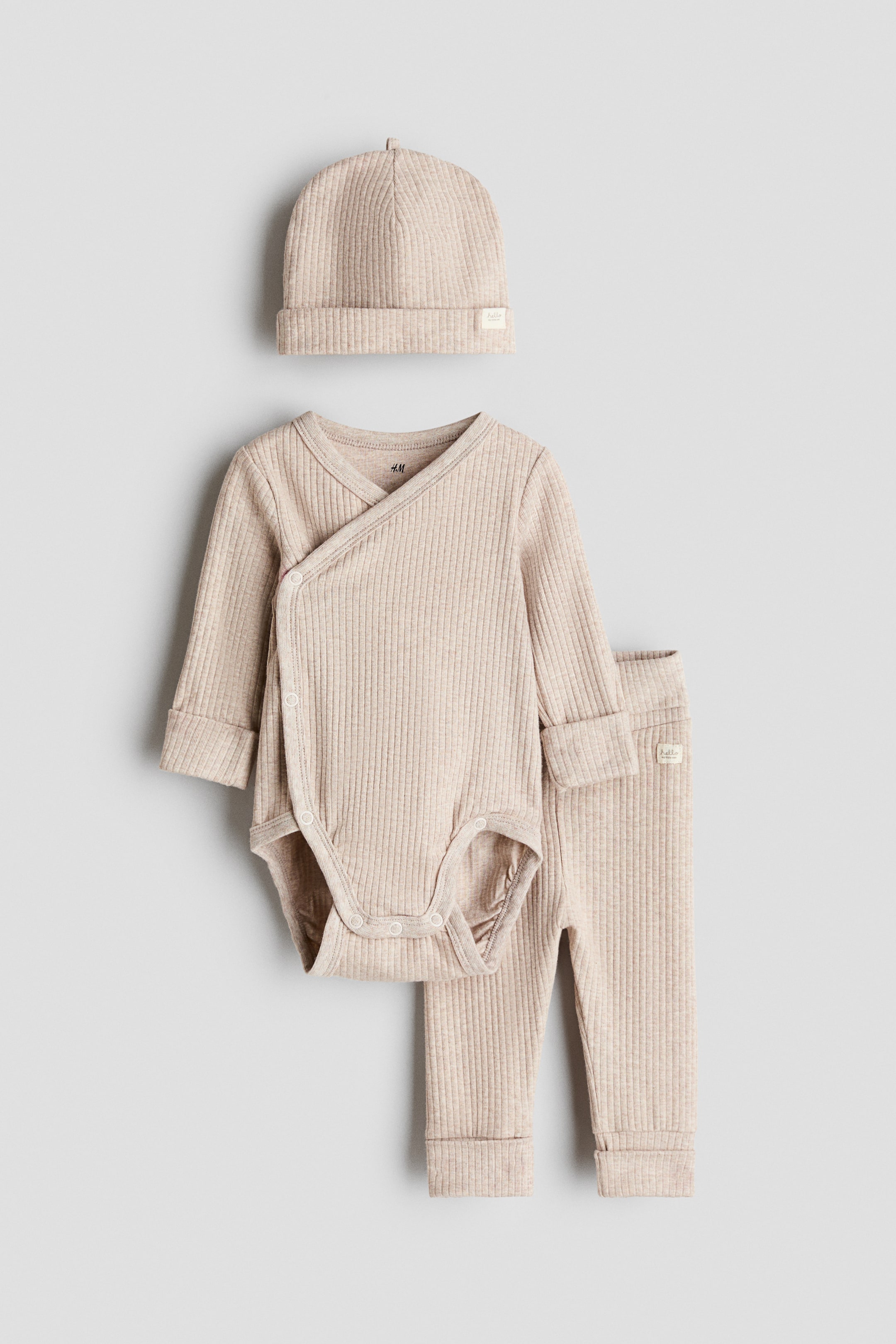 Agrandir l'image: Un ensemble bébé trois pièces en maille côtelée beige clair est présenté de face, comprenant un body croisé à manches longues avec fermeture à boutons-pression diagonale et revers aux poignets, un pantalon assorti avec revers aux chevilles, ainsi qu’un bonnet à bord relevé.