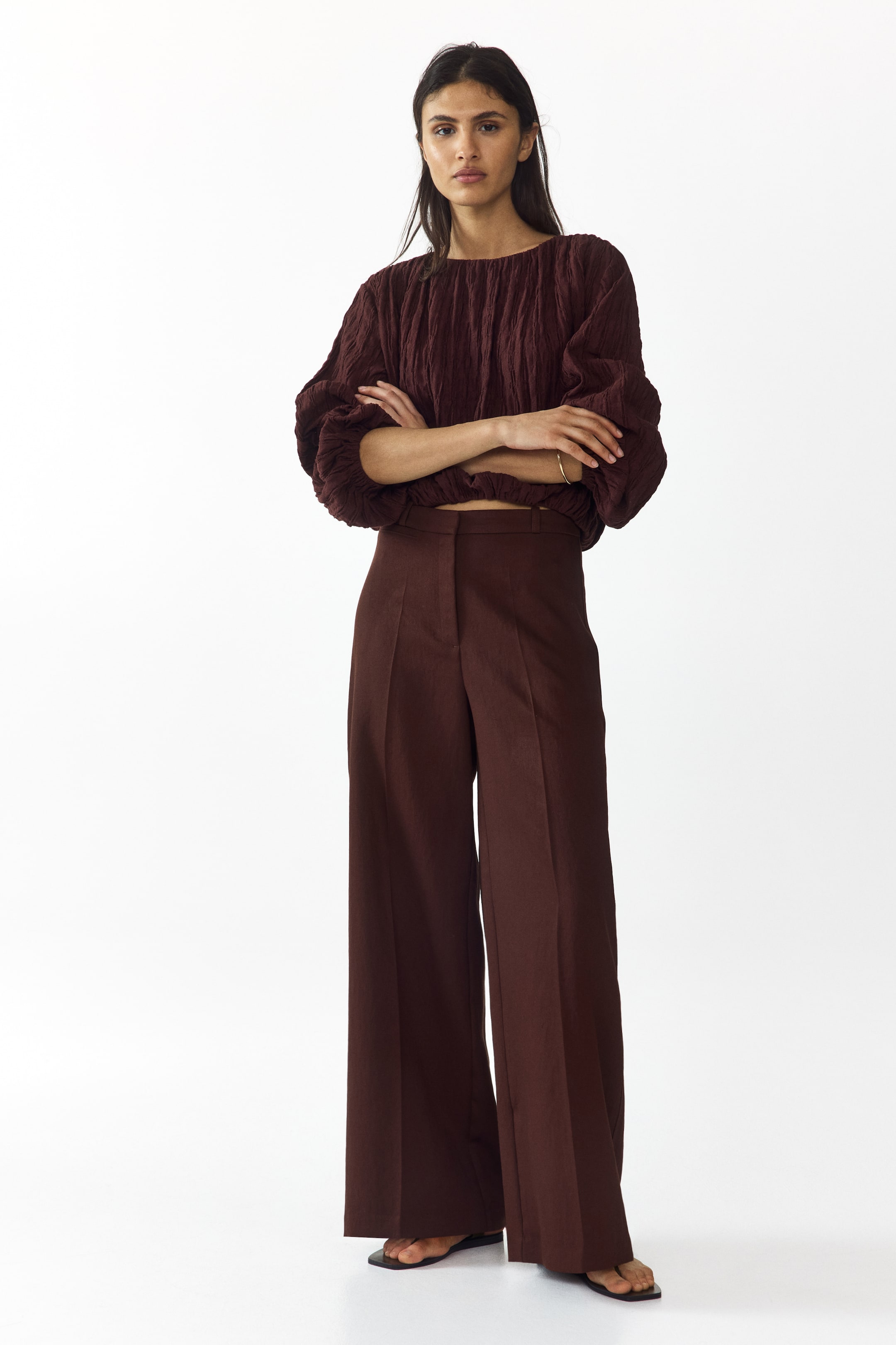 View larger image: A dark brown crinkled top is styled with a round neckline, voluminous long sleeves with gathered cuffs, and a gathered cropped hem. It pairs perfectly with high-waisted, wide-leg dark brown trousers featuring front creases, elegantly covering black open-toed sandals.