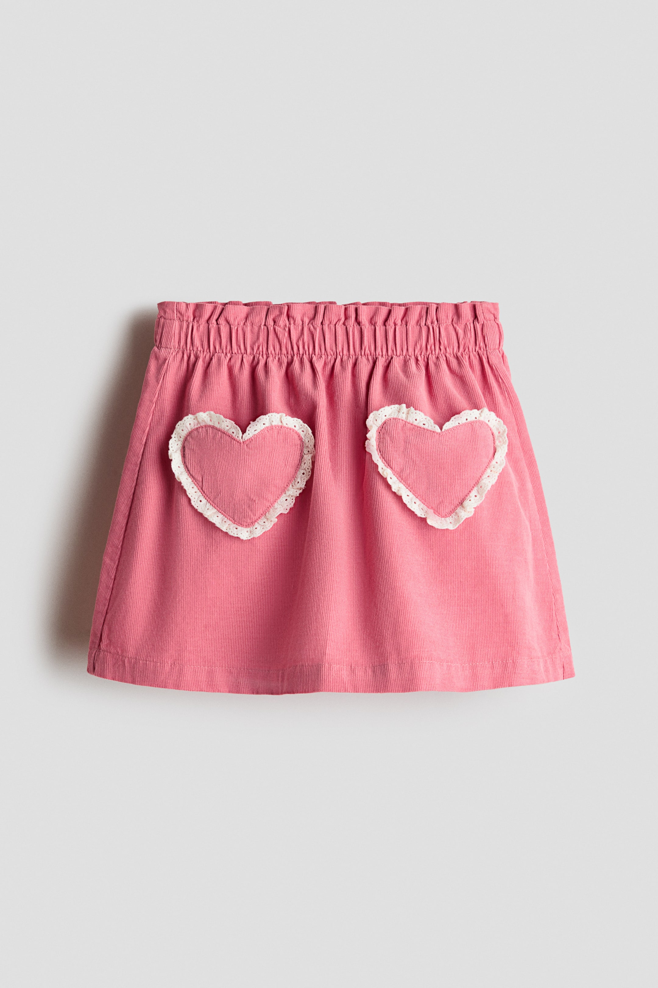 View larger image: A pink corduroy skirt features a shirred elastic waistband and two heart-shaped patch pockets on the front, finished with white lace trim.