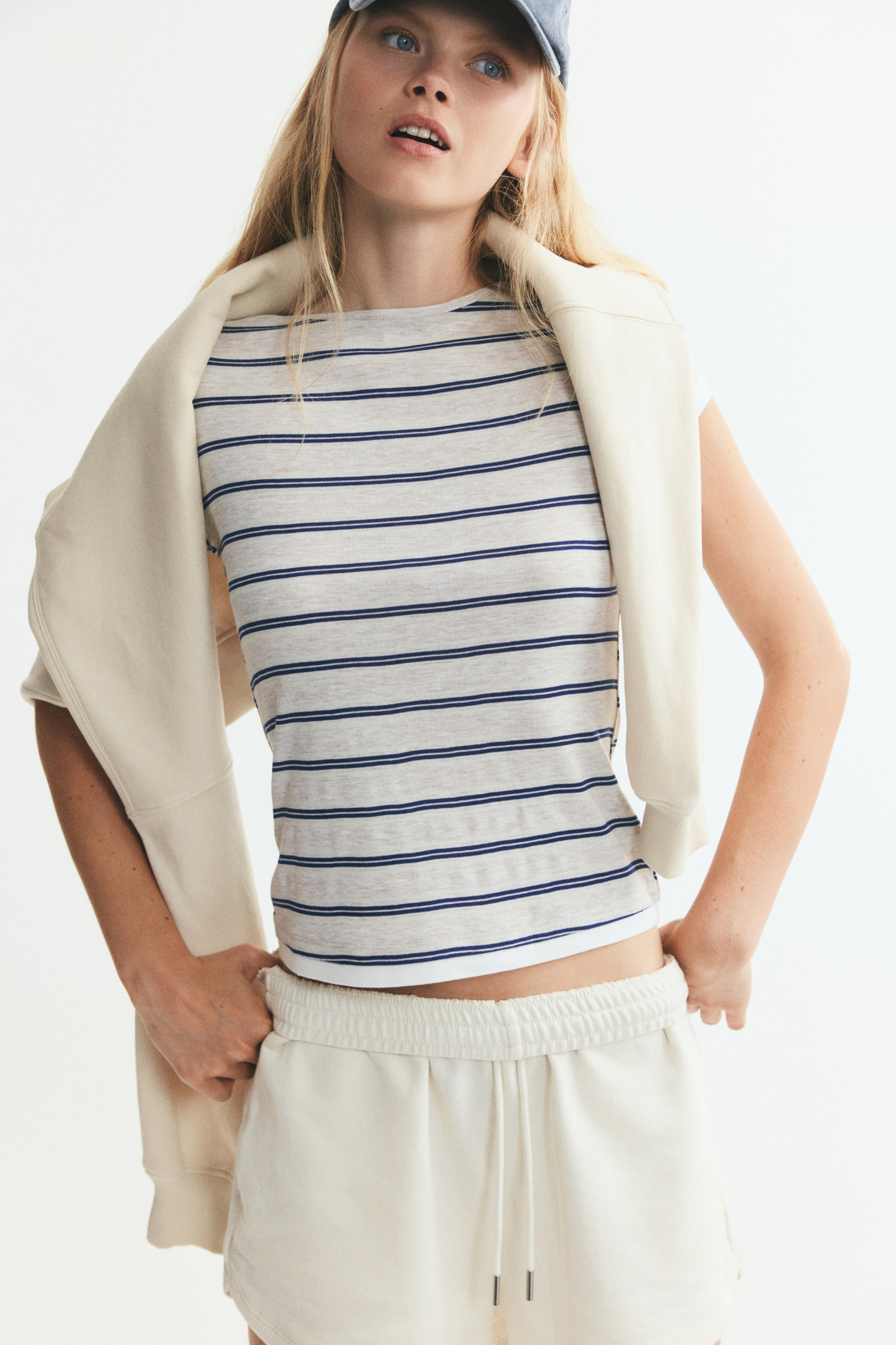 View larger image: A short, fitted T-shirt in striped jersey is being worn by the model, featuring a round neckline and a contrasting white trim at the hem. The model is styling the T-shirt with cream-coloured drawstring shorts and a matching sweatshirt draped over the shoulders.