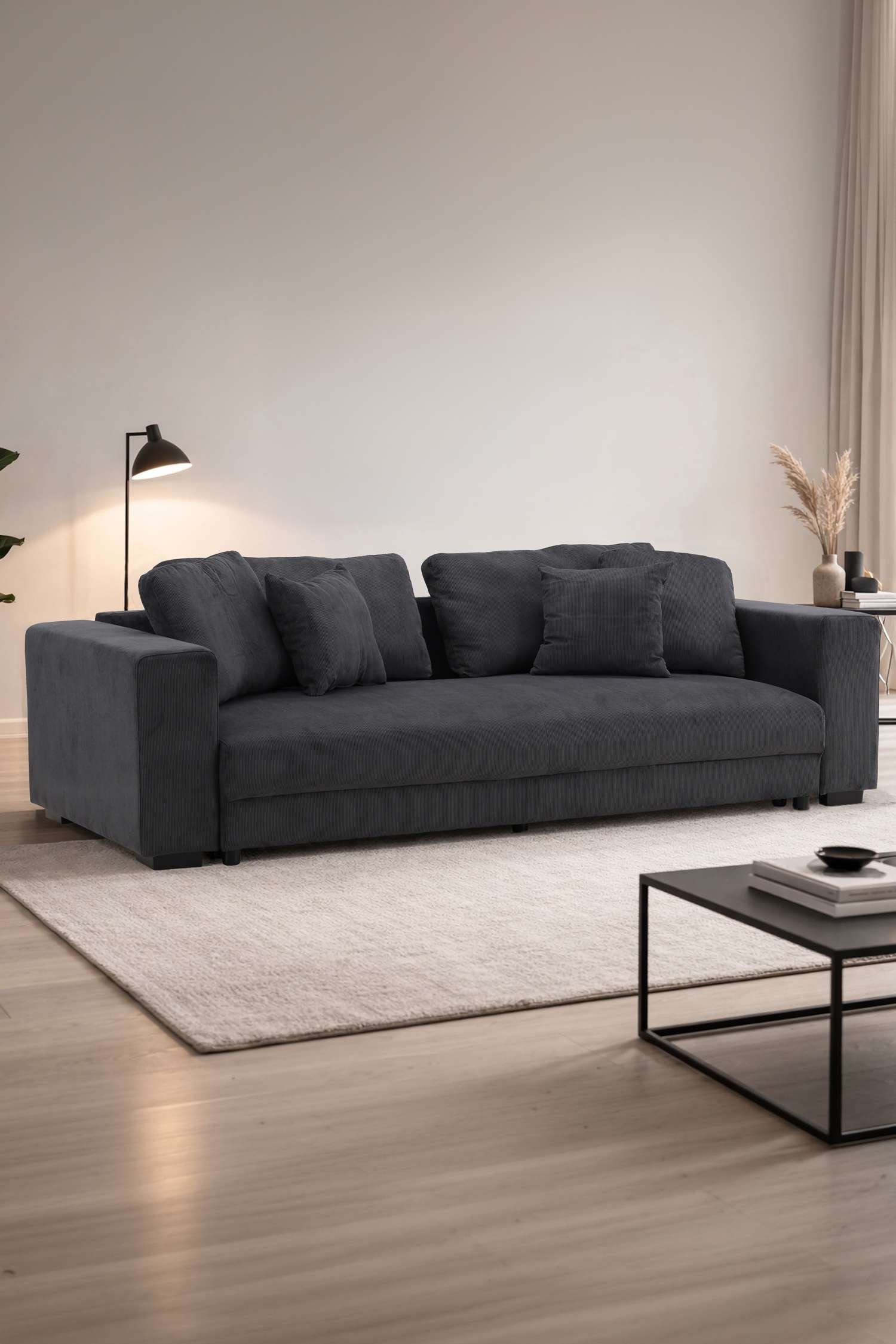Een donkergrijze sofa staat naar voren gericht, met een rechthoekige vorm en brede, rechte armleuningen. De sofa heeft meerdere rugkussens en kleinere sierkussens, allemaal bekleed met een geribbelde stof.