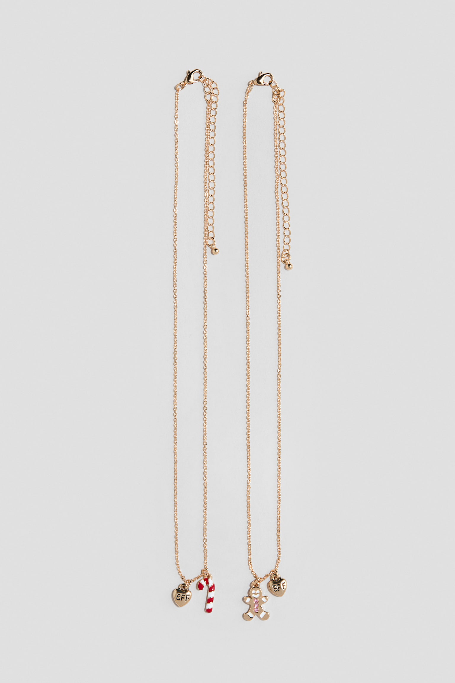 Dos collares de cadena en tono dorado cuelgan verticalmente, con charms festivos en la parte inferior. El collar de la izquierda lleva un charm de corazÃ³n en tono dorado grabado con Â«BFFÂ» y un charm de bastÃ³n de caramelo a rayas rojas y blancas, mientras que el collar de la derecha lleva un charm de corazÃ³n Â«BFFÂ» a juego y un charm de hombre de jengibre con detalles de gema rosa. Ambas piezas muestran cierres ajustables de mosquetÃ³n con cadenas de extensiÃ³n en la parte superior.
