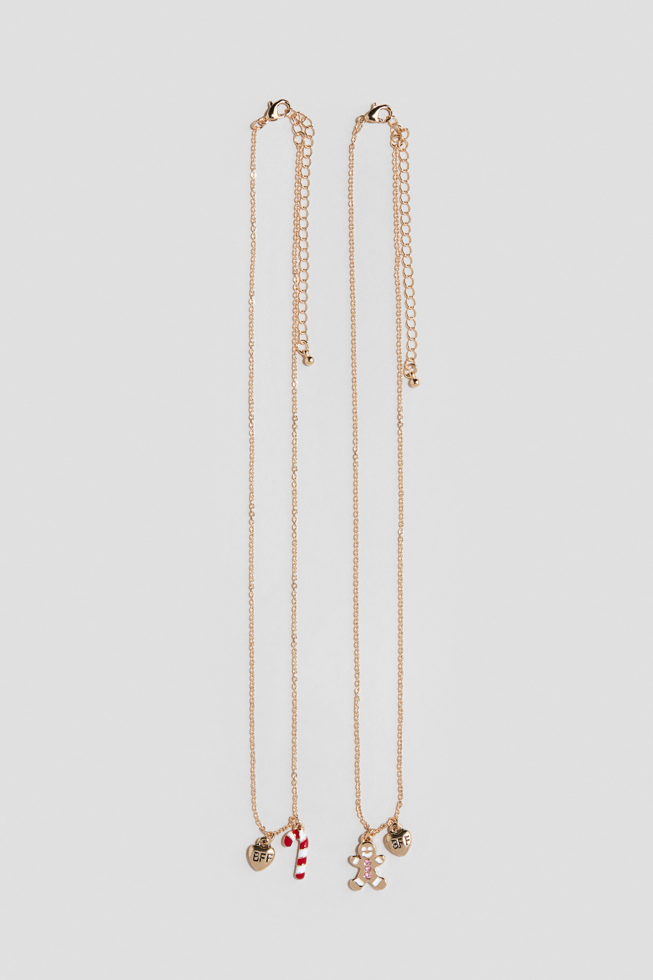 Ver imagen más grande: Dos collares de cadena en tono dorado cuelgan verticalmente, con charms festivos en la parte inferior. El collar de la izquierda lleva un charm de corazón en tono dorado grabado con «BFF» y un charm de bastón de caramelo a rayas rojas y blancas, mientras que el collar de la derecha lleva un charm de corazón «BFF» a juego y un charm de hombre de jengibre con detalles de gema rosa. Ambas piezas muestran cierres ajustables de mosquetón con cadenas de extensión en la parte superior.