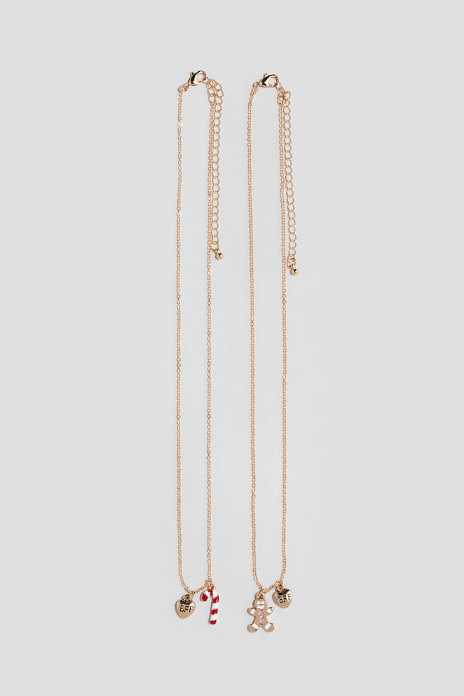 Two gold-toned chain necklaces are hanging vertically, featuring festive charms at the bottom. The left necklace is holding a gold-toned heart charm engraved with "BFF" and a red-and-white striped candy cane charm, while the right necklace is holding a matching "BFF" heart charm and a gingerbread man charm with pink gemstone details. Both pieces are showing adjustable lobster clasp closures with extender chains at the top. - 1