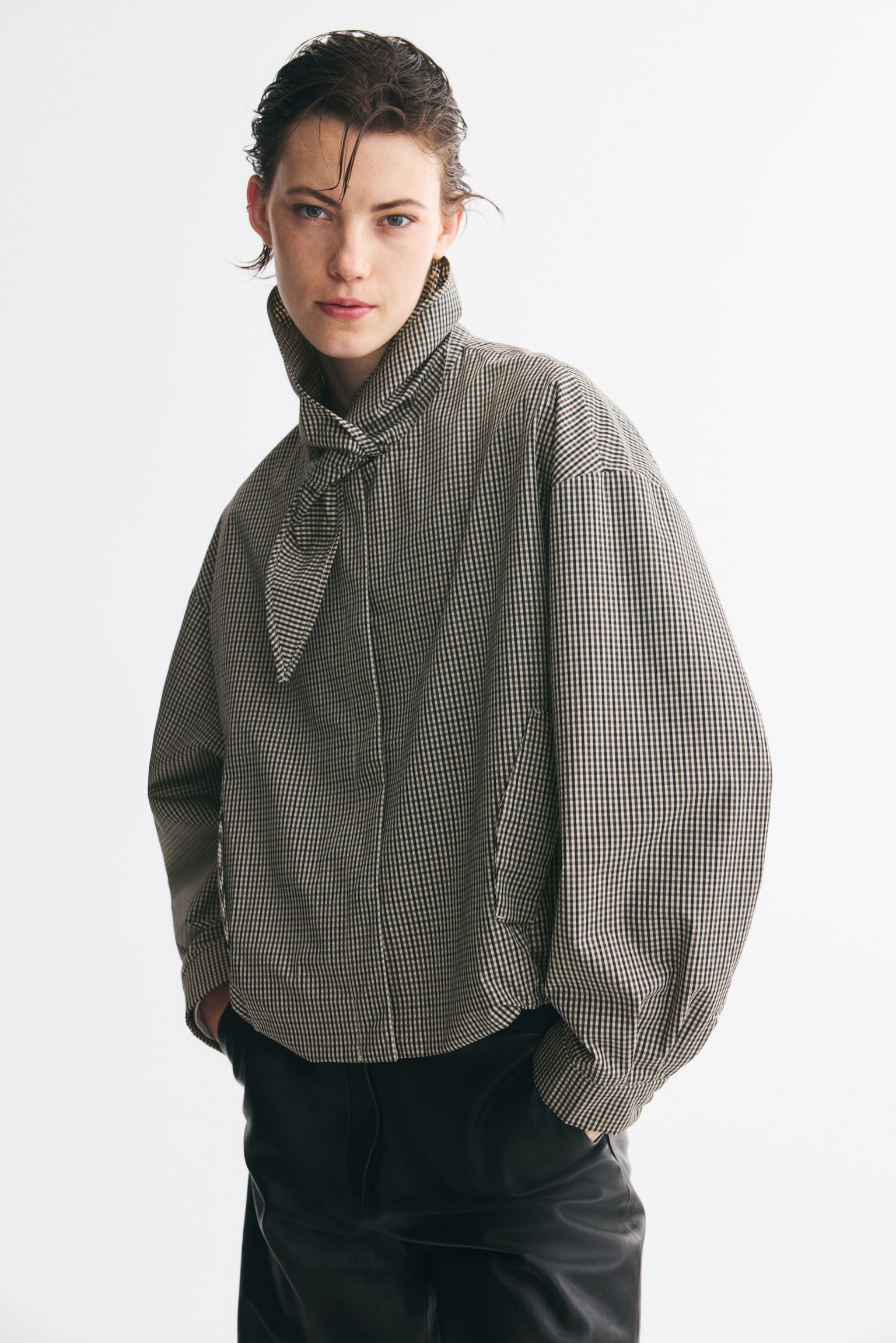 A cropped jacket is displaying a black and off-white gingham pattern, with a large pointed collar tying into a bow at the front. It is featuring two angled patch pockets, long sleeves with buttoned cuffs, and a gathered bottom hem. - 1