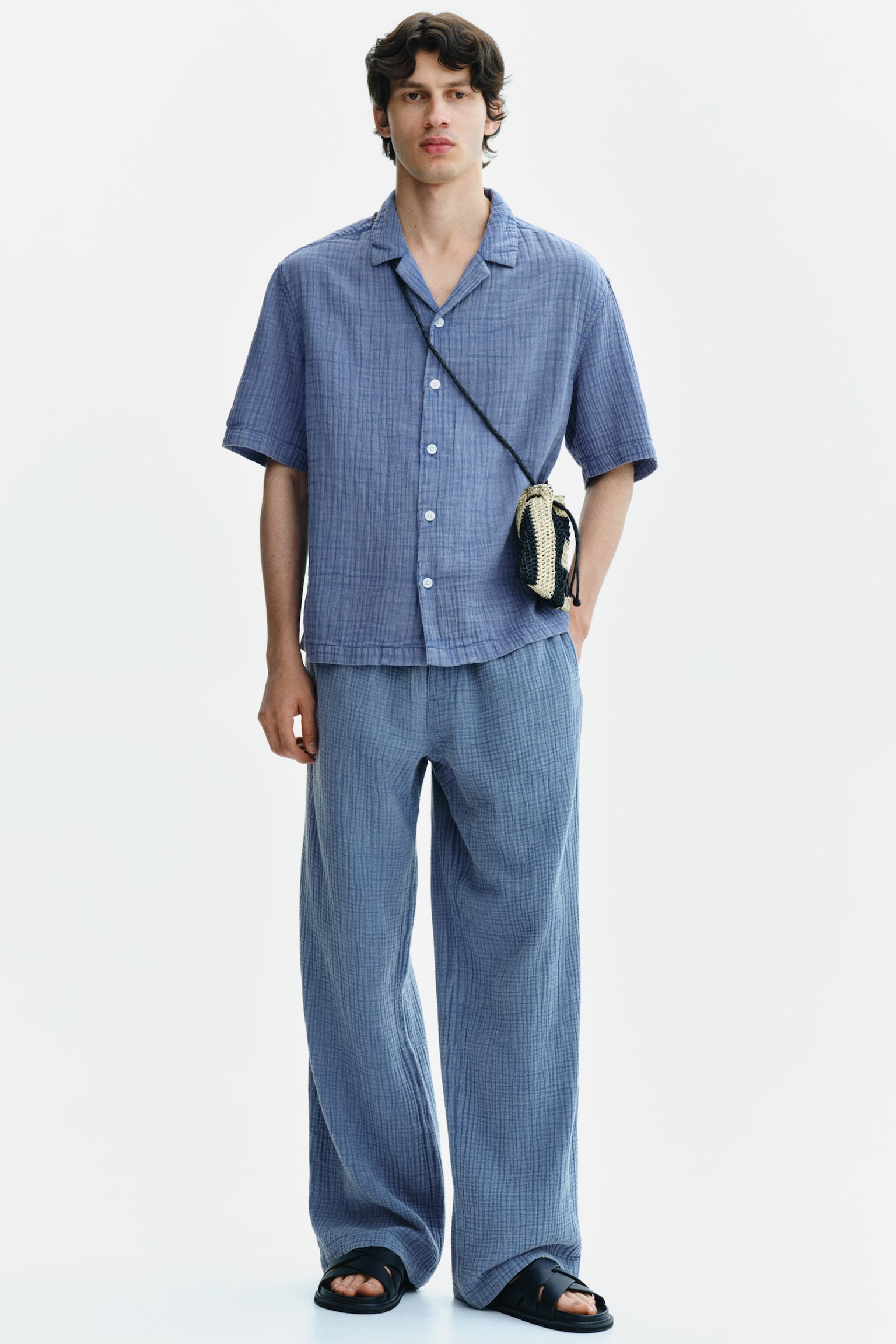 The model is wearing trousers crafted from cotton muslin, boasting an elasticated waist, a relaxed fit, and a roomy silhouette. One hand is casually slipped into a subtle side pocket. - 1