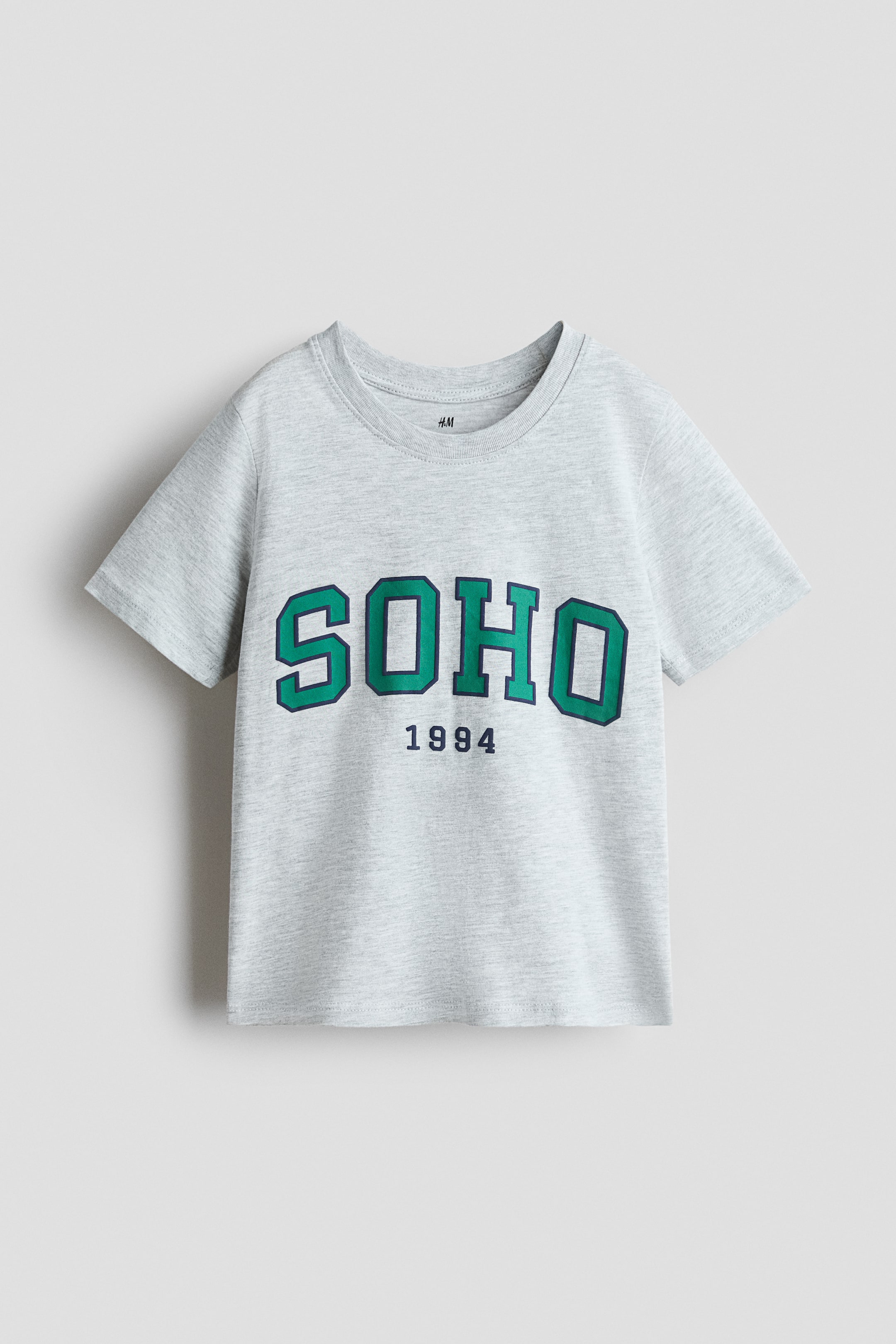 Nagyobb kép megtekintése: Egy világosszürke melírozott rövid ujjú póló elölről látható, kerek nyakkivágással és H&M márkajelzéssel a belső hátsó nyakrészén. A póló mellrészén grafikai nyomat található: a "SOHO" nagy, zöld blokkbetűkkel, sötétkék kontúrral körvonalazva, alatta pedig kisebb, sötétkék "1994" felirat.