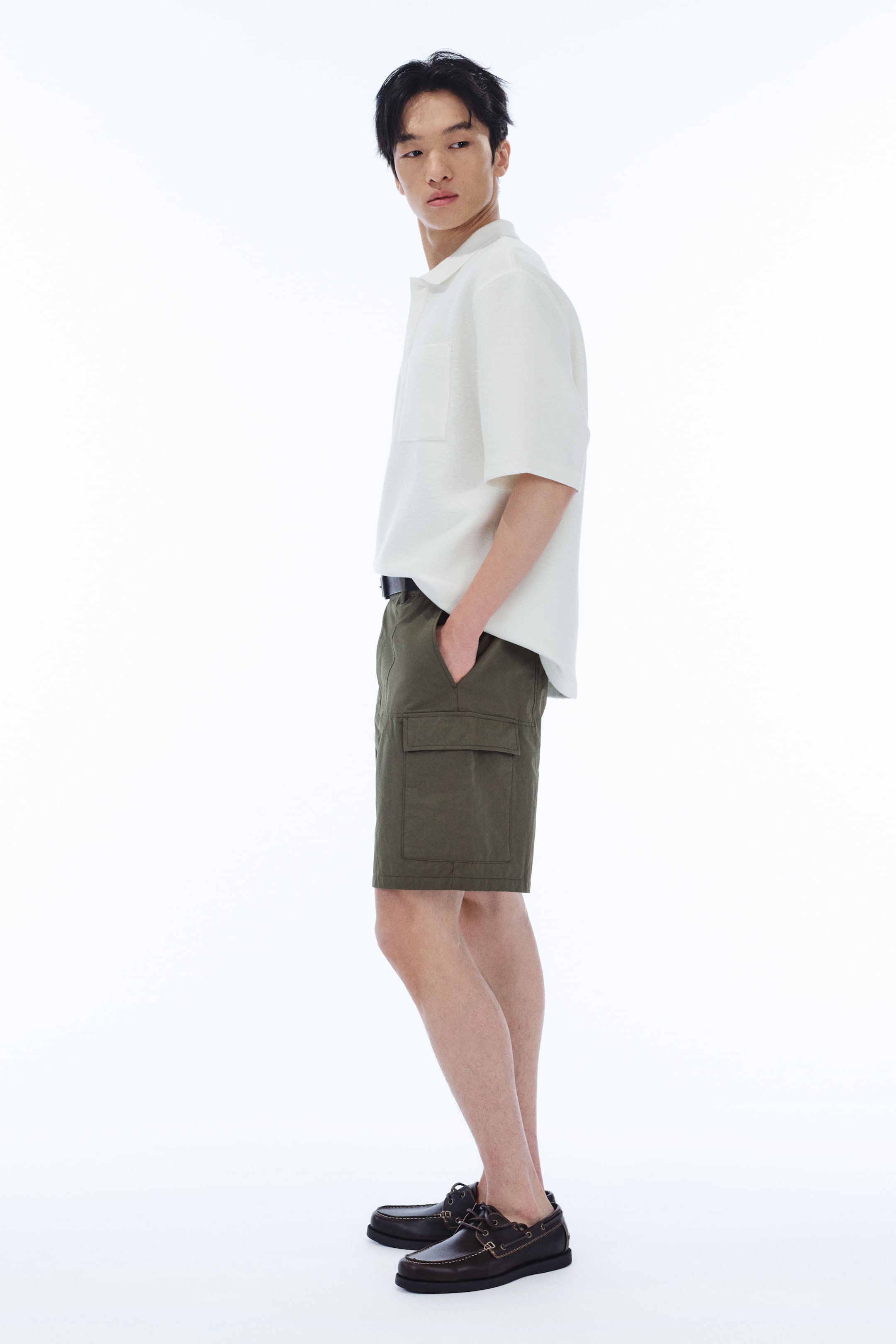 View larger image: The model is wearing olive green cargo shorts with a relaxed fit, showcasing diagonal side pockets and patch leg pockets with flaps. One hand is casually placed in a diagonal side pocket. The ensemble is completed with a white shirt, black belt, and brown boat shoes.