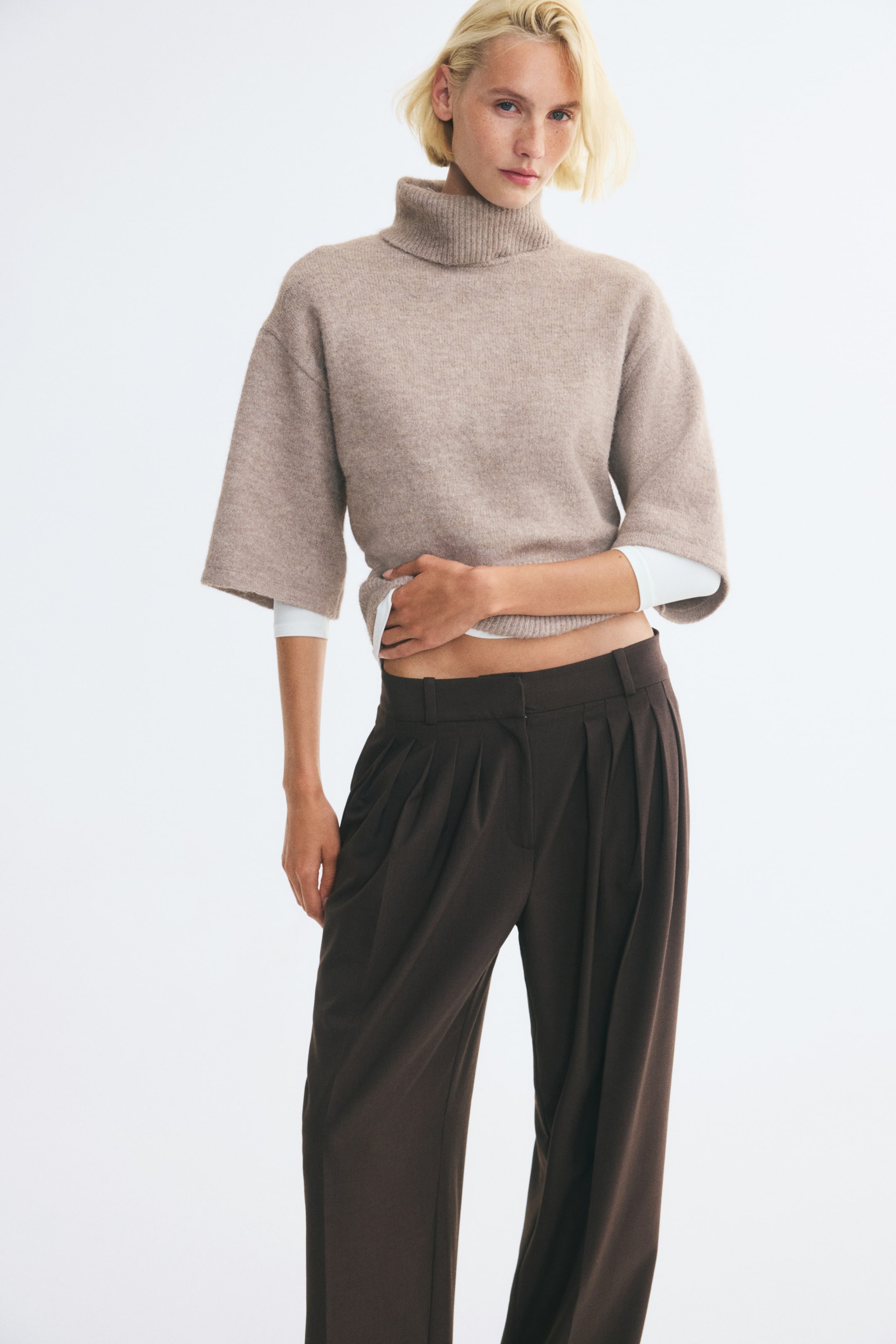 View larger image: A beige knitted sweater featuring a ribbed high neck, dropped shoulders, and wide elbow-length sleeves is being worn by the model over a white long-sleeved top. The model is holding the ribbed hem of the sweater with one hand, styling it with dark brown trousers.