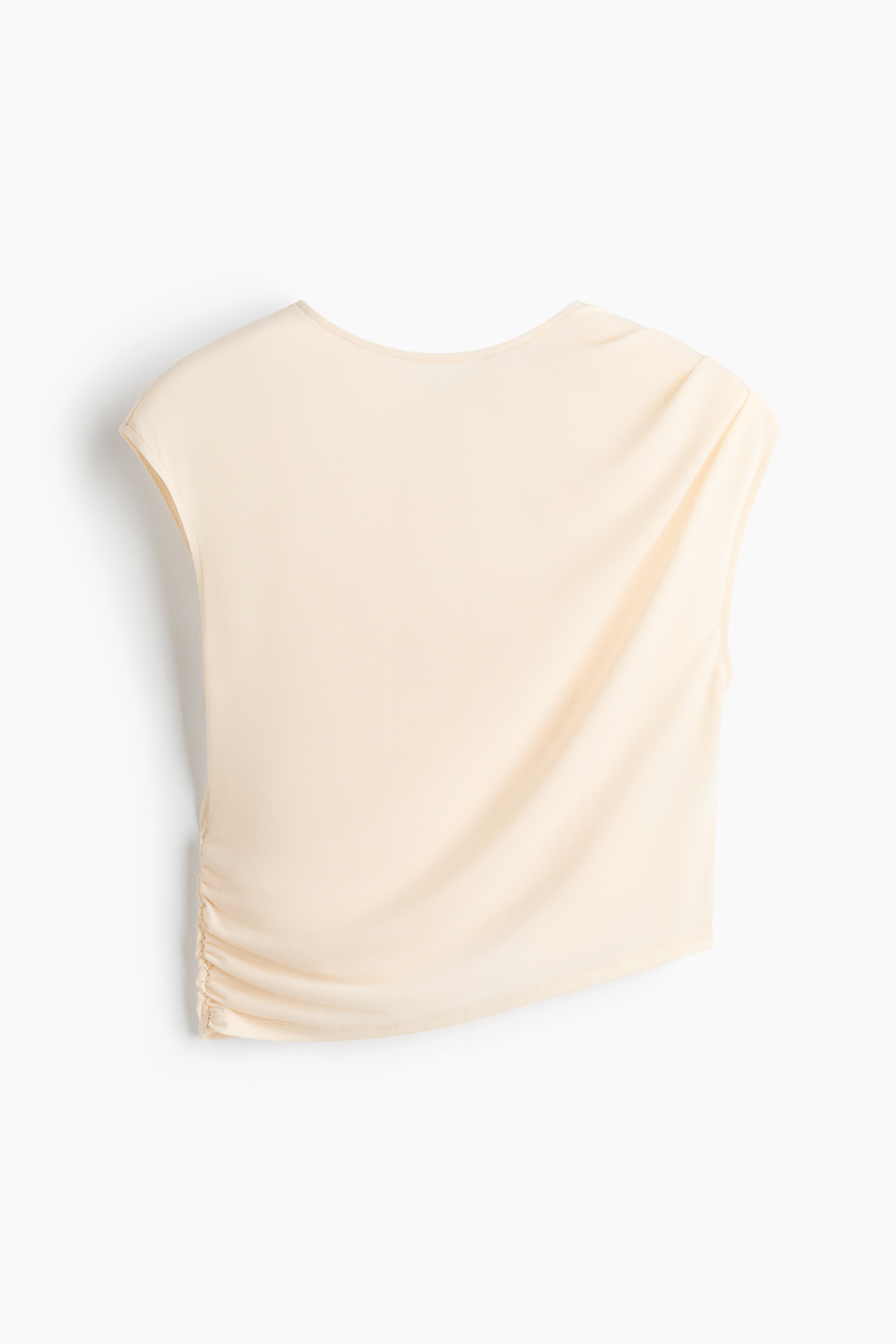 A cream-coloured jersey top featuring a round neckline, cap sleeves, and draped pleating on one shoulder is being worn by the model. The cropped top is styled with a matching gathered skirt and features a gathered side seam that creates a gently draped, asymmetrical hem.