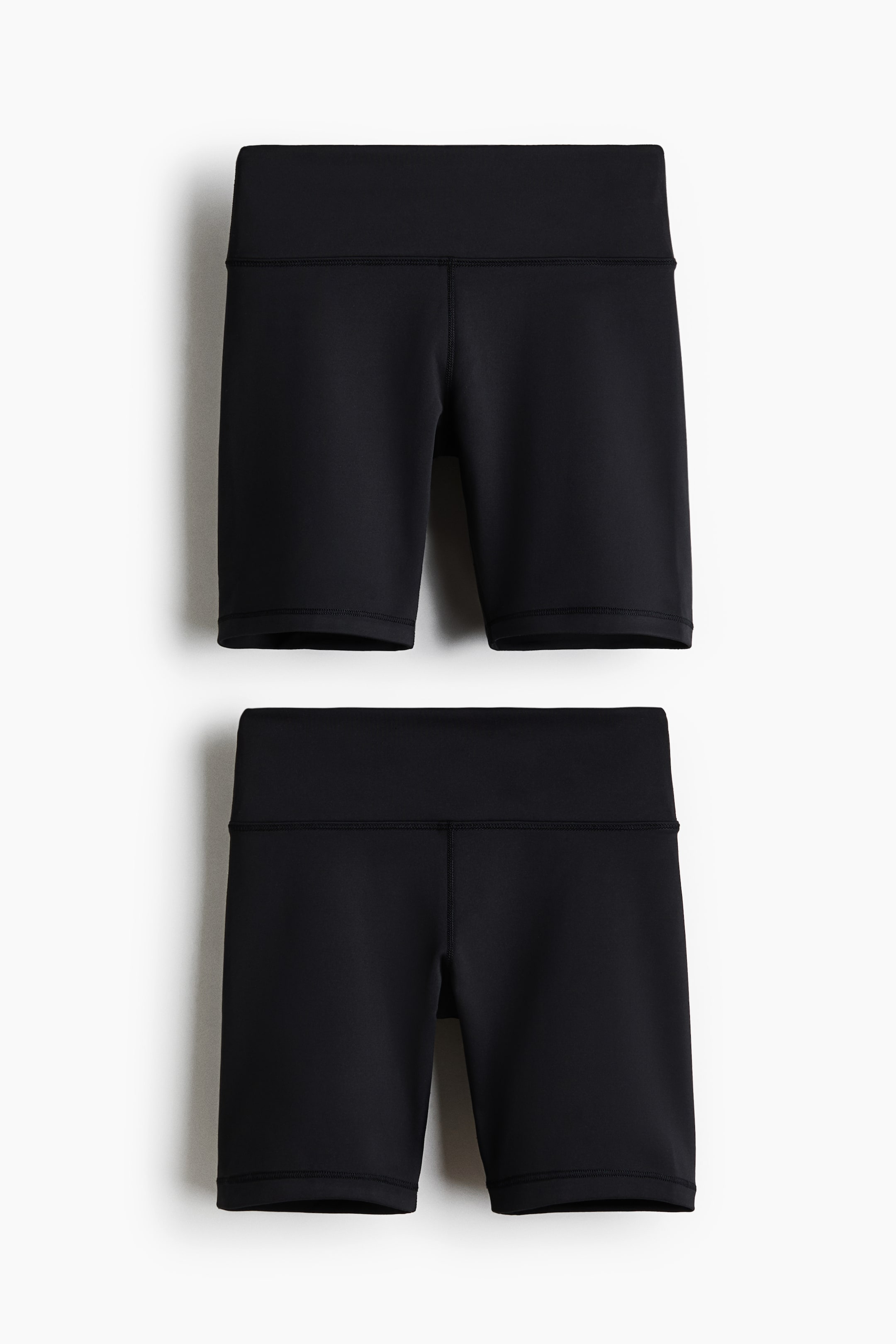 View larger image: Two pairs of black cycling shorts are shown, each made from smooth, stretchy fabric with a wide, high-rise waistband and visible flatlock seams along the legs and hem.