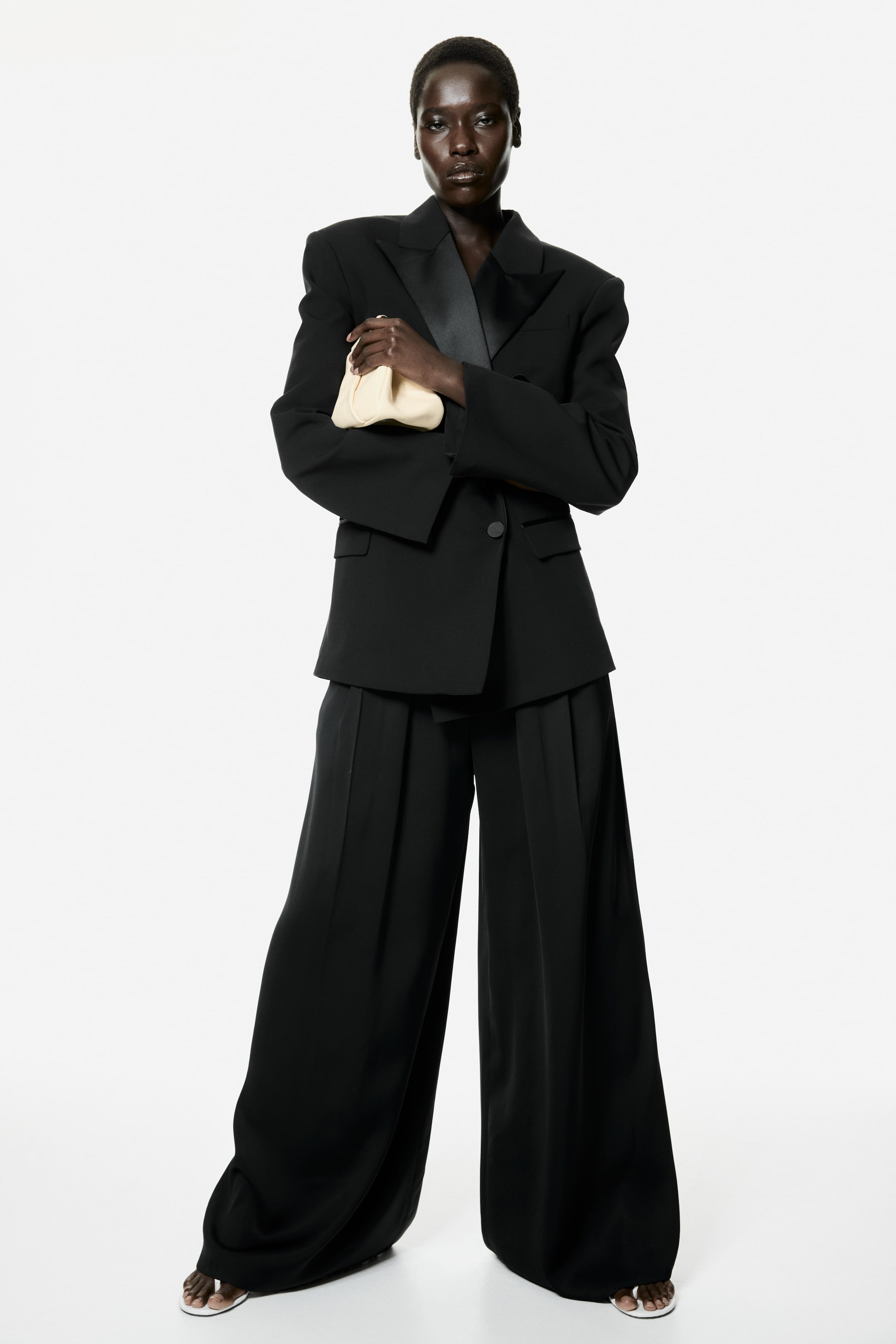View larger image: The model is showcasing loose-fit black trousers, featuring wide legs with pleated panels at the top, elegantly draping over white sandals.