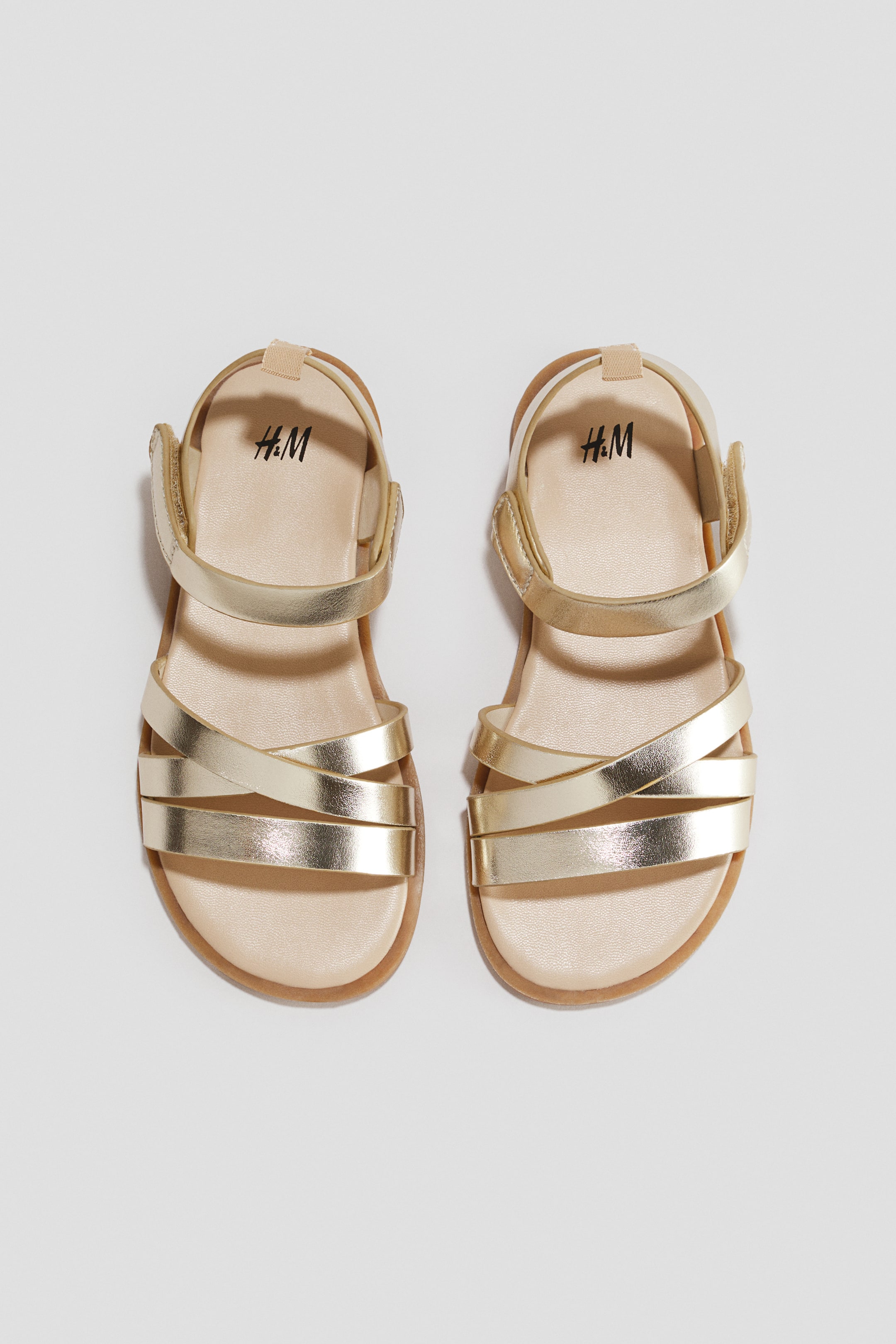 View larger image: A pair of gold metallic sandals is resting, viewed from an overhead front angle, featuring three front straps with two crisscrossing over the instep. An adjustable ankle strap with a hook and loop fastener is securing the heel, complementing the beige footbed and thin tan sole.