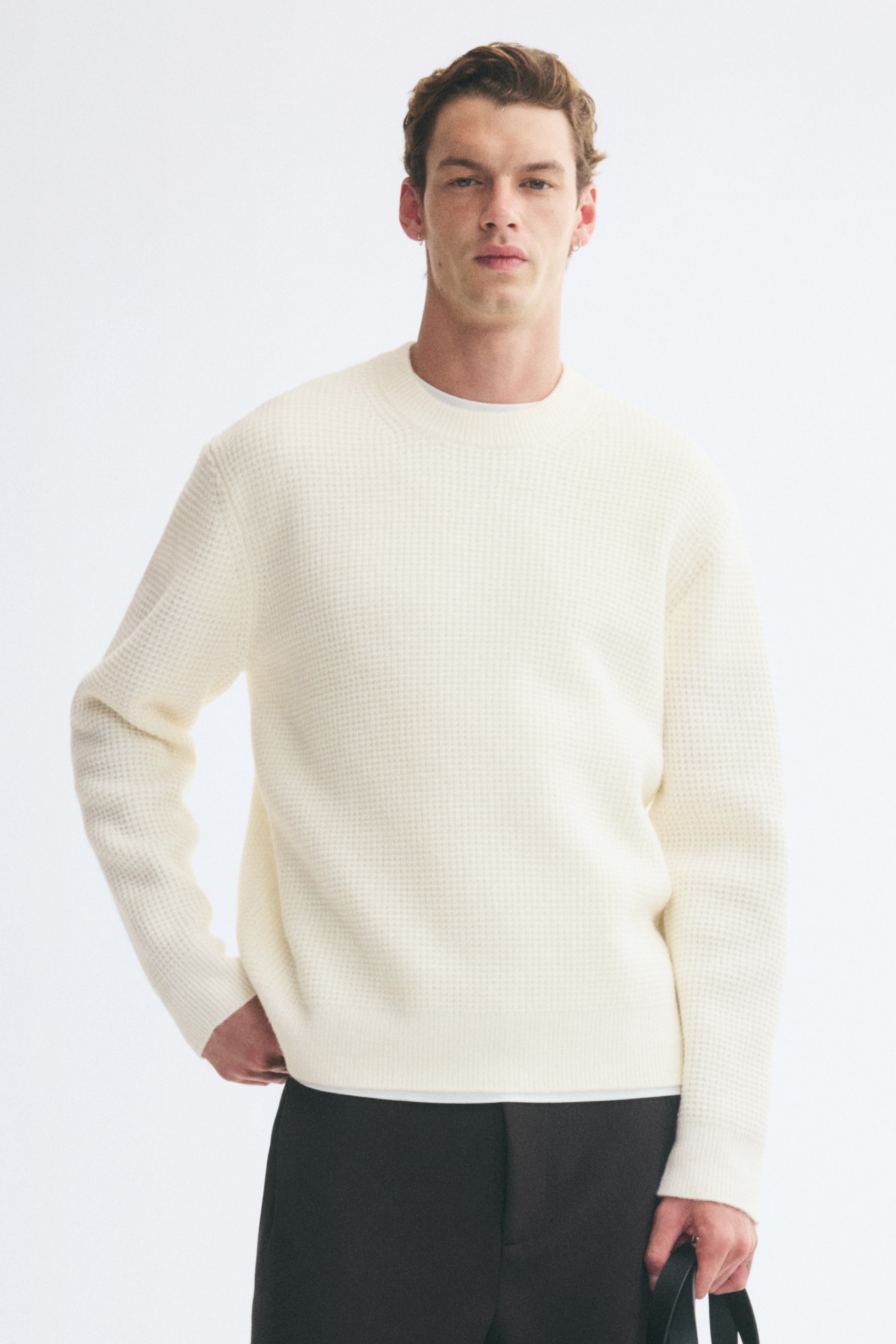 Größeres Bild ansehen: Ein strukturierter cremefarbener Strickpullover mit rundem, geripptem Ausschnitt, gerippten Bündchen und geripptem Saum wird vom Model getragen. Der Pullover in lockerer Passform wird über einem weißen T-Shirt getragen und mit dunklen Hosen kombiniert.