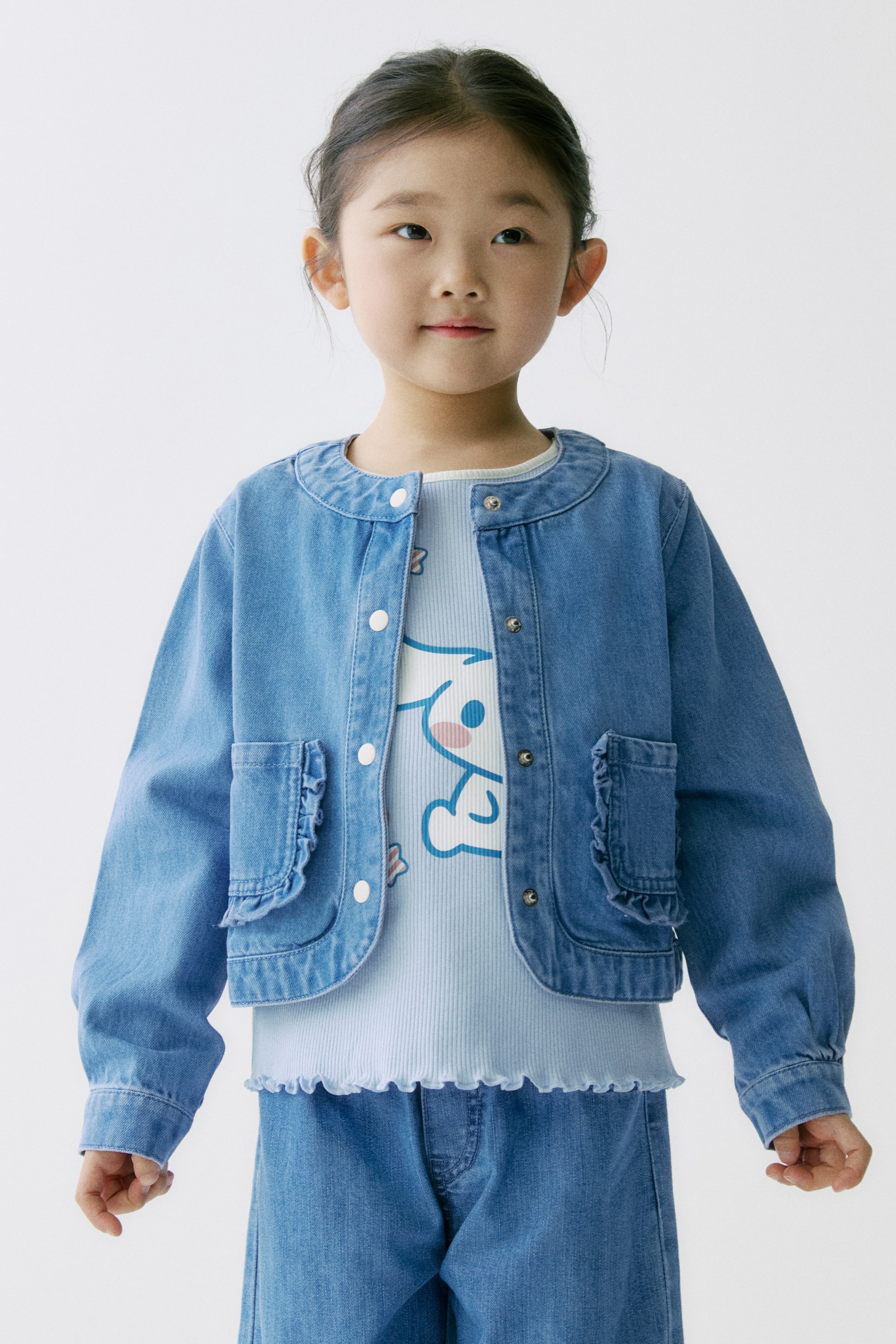 拡大画像を表示: ライトブルーのデニムジャケットは、ノーカラーのラウンドネックと白いスナップボタンのフロント開閉を備えています。前面にはフリル付きのパッチポケットが2つ施され、ボタンカフス付きの長袖で、ライトブルーのリブトップの上に前を開けて羽織るように着用されています。