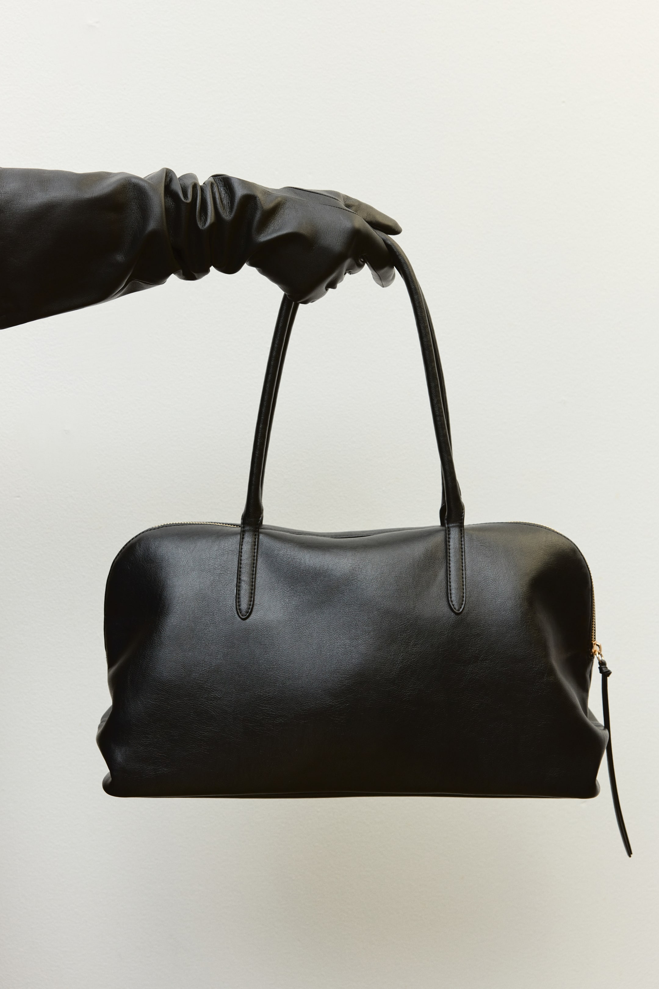 Größeres Bild ansehen: Eine schwarze Lederhandtasche mit abgerundeter Silhouette und schmalen, doppelten Schulterriemen wird von dem Model getragen, das einen langen schwarzen Lederhandschuh trägt. Die Handtasche verfügt über einen Reißverschluss oben mit einer langen Leder-Zuglasche.
