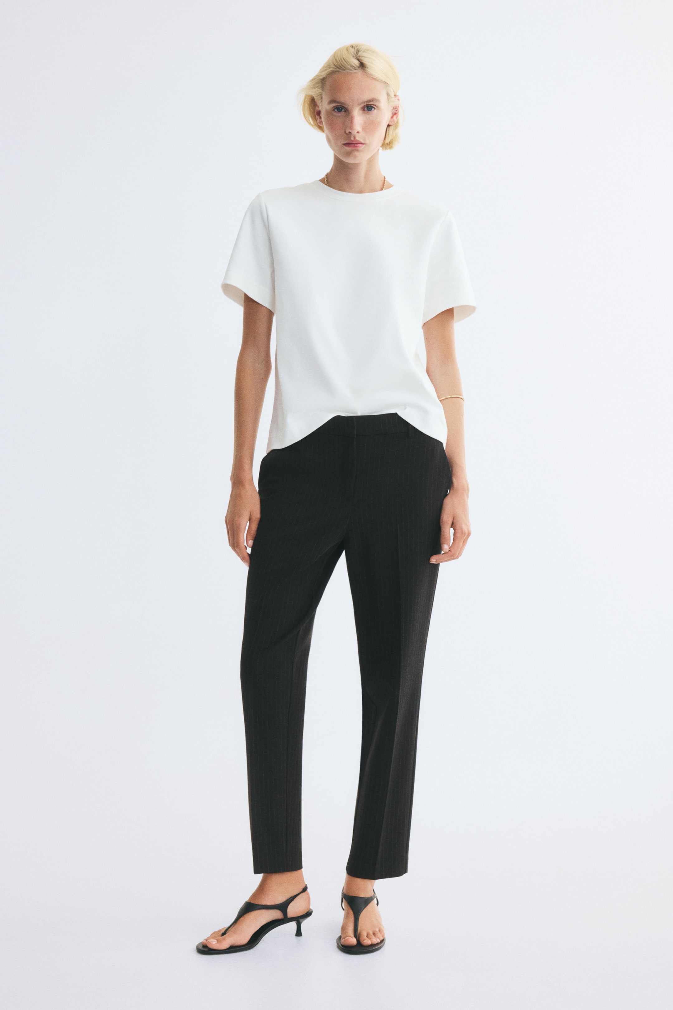 Grotere afbeelding bekijken: Zwarte cigarette-broek tot op de enkel, met taps toelopende pijpen en een subtiel verticaal patroon, gedragen door het model. De broek is gestyled met een wit T-shirt met korte mouwen en zwarte sandalen met hak en teenlus.