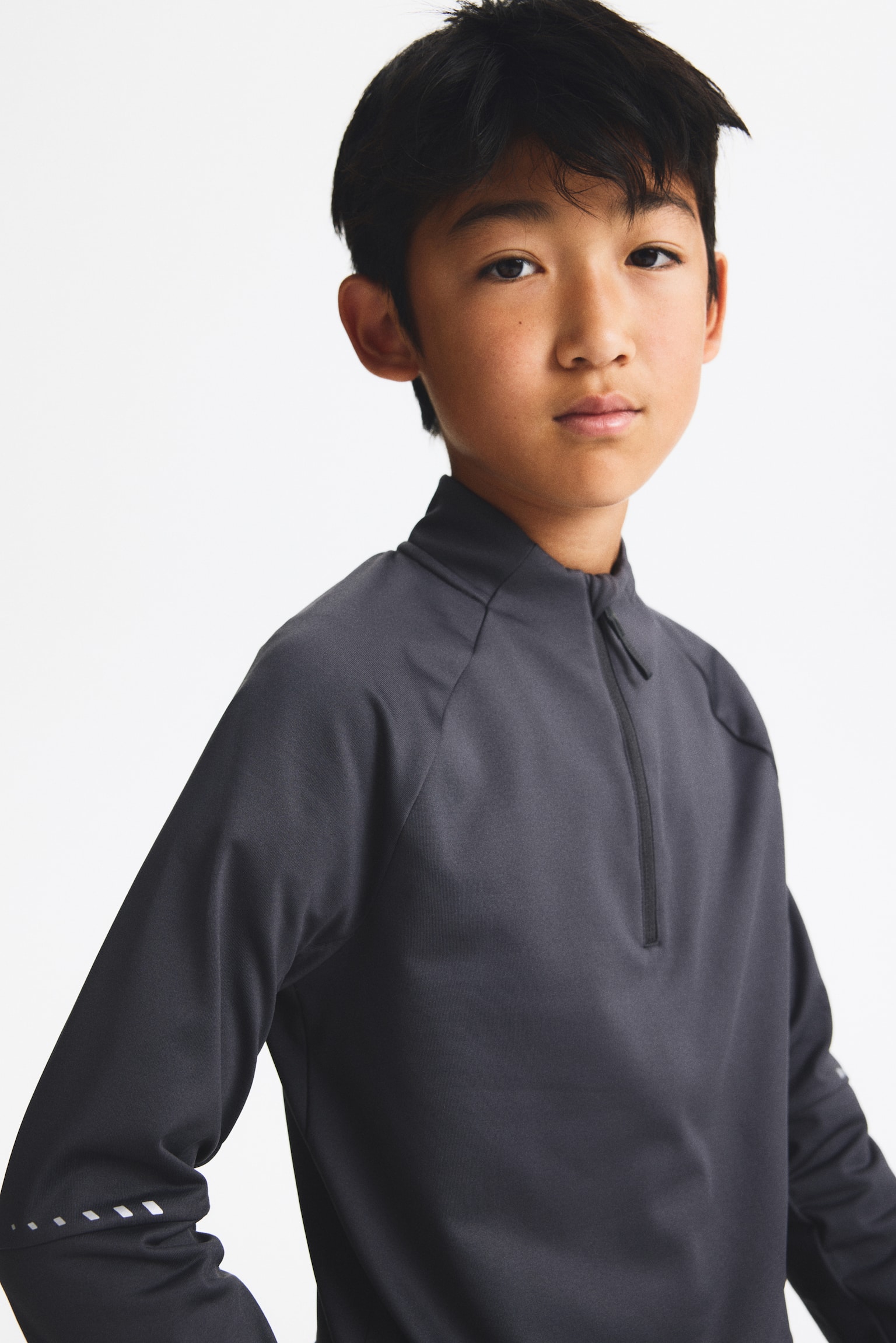 A dark grey long-sleeved athletic top is being worn front-facing, featuring a high collar with a quarter-zip fastening and raglan sleeves. The top also features small reflective dashed details on the lower sleeve near the cuff. - 1