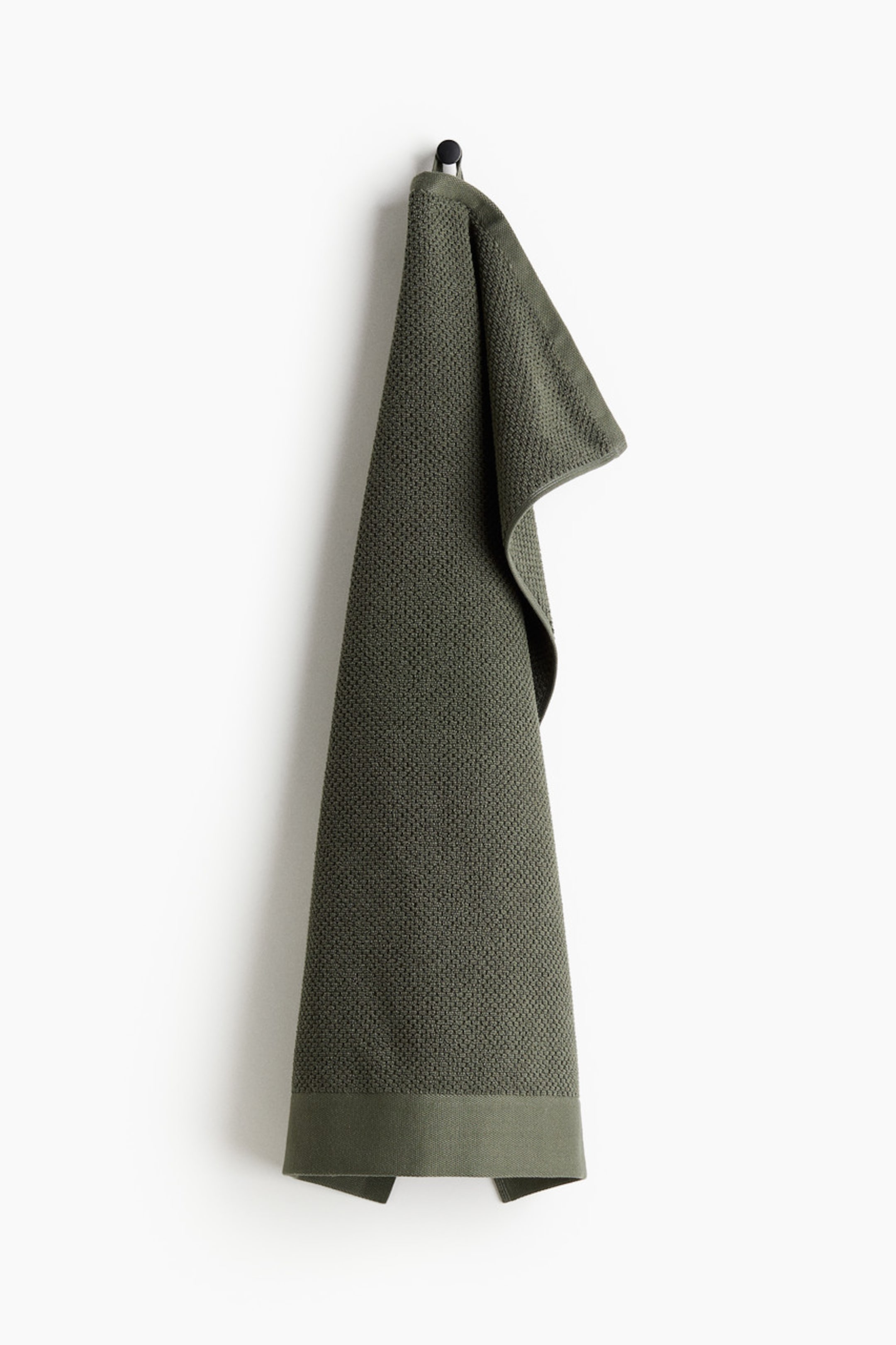 View larger image: An olive green towel is hanging folded over a black hook. It showcases a waffle weave texture throughout and a smooth, plain woven band at its bottom edge.