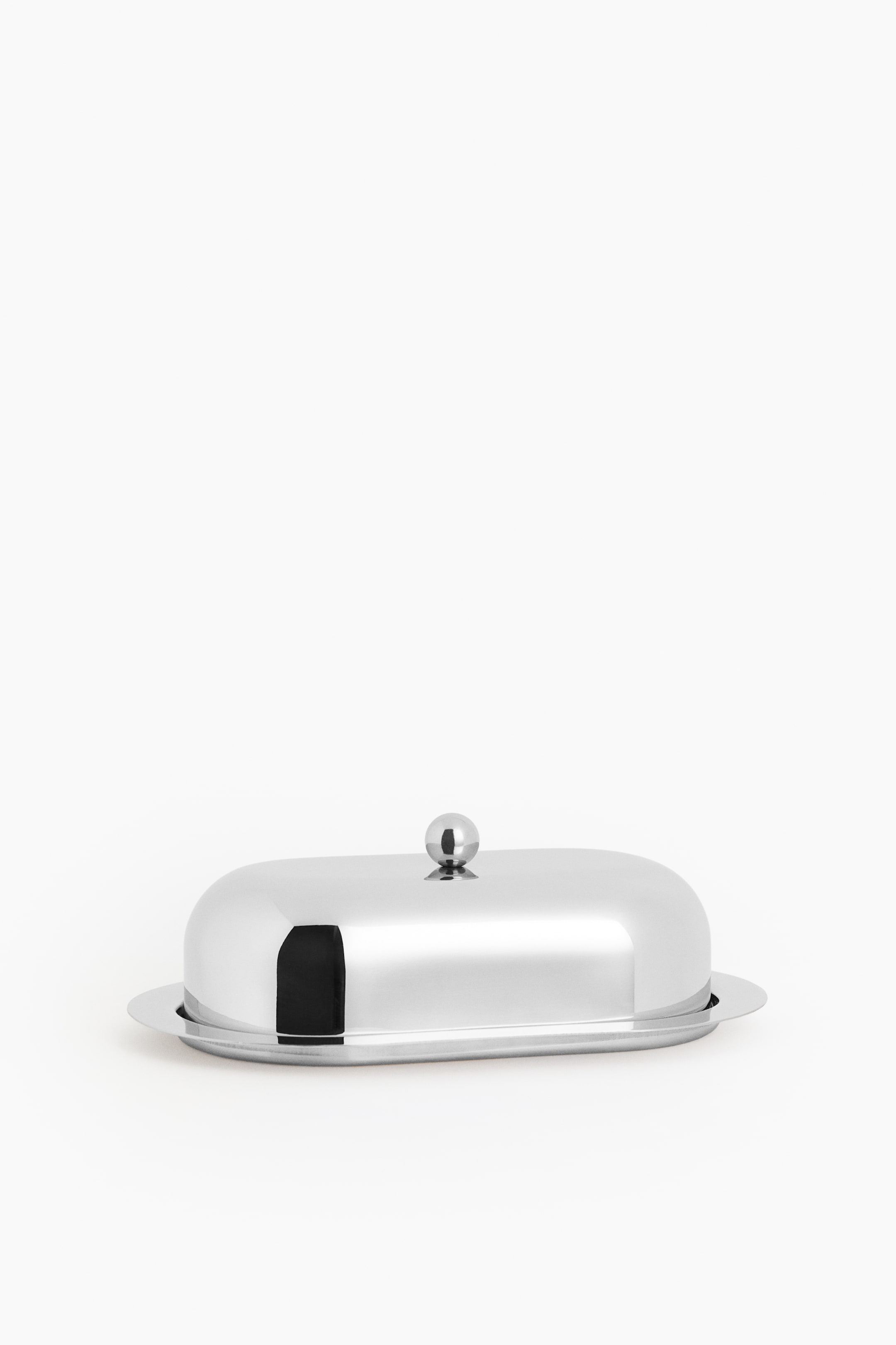 View larger image: The butter dish is positioned front-facing, highlighting its sleek stainless steel build and oval shape. It features a cloche lid with a small round knob on top, set on an oval tray.